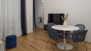 A modern living room with a round white table, dark blue chairs, a small potted plant, and a flat-screen TV on the wall.