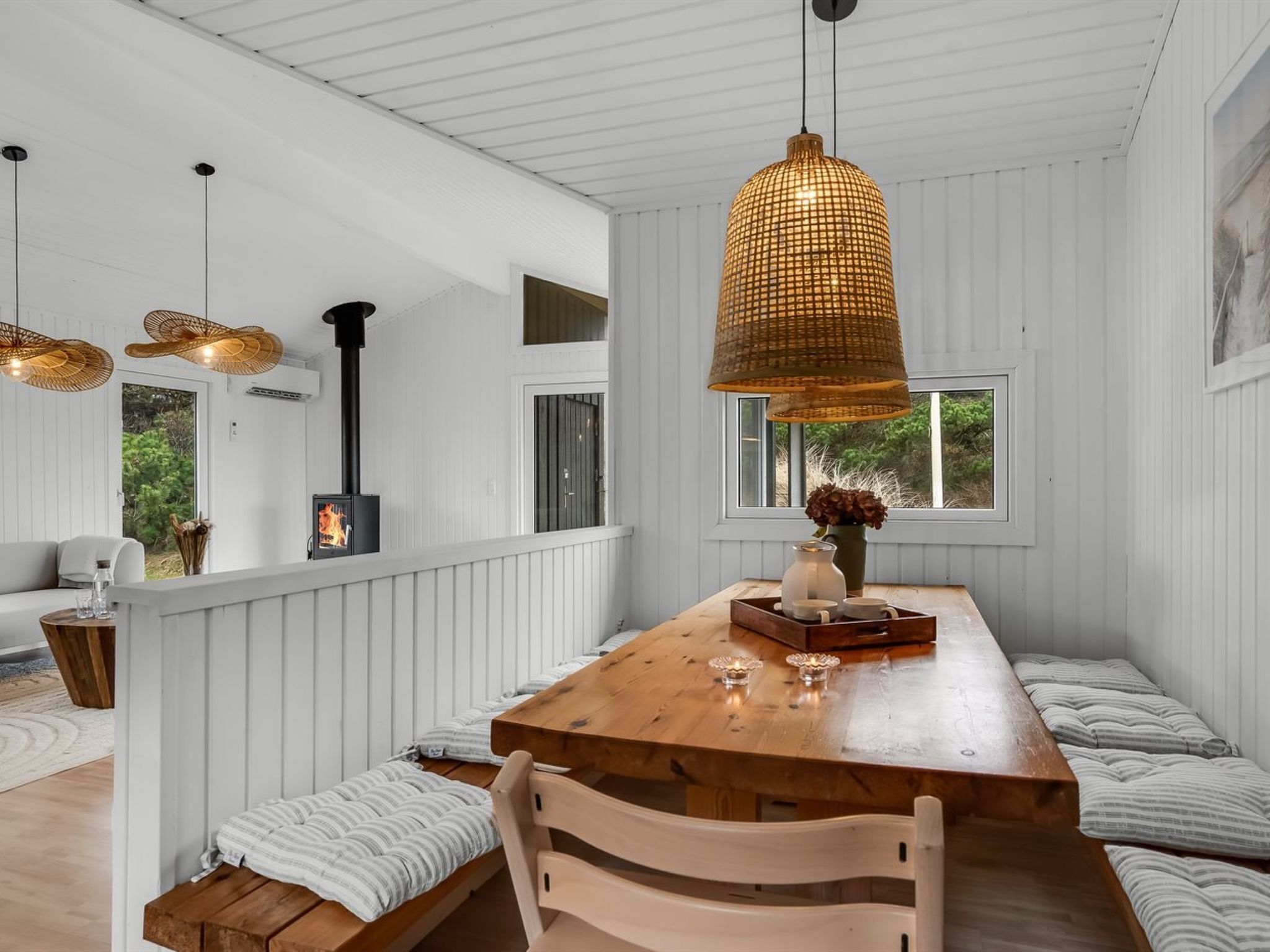 Ein gemütliches Interieur mit einem Holztisch, Sitzbänken, Korbtreleuchtern und Blick ins Grüne.