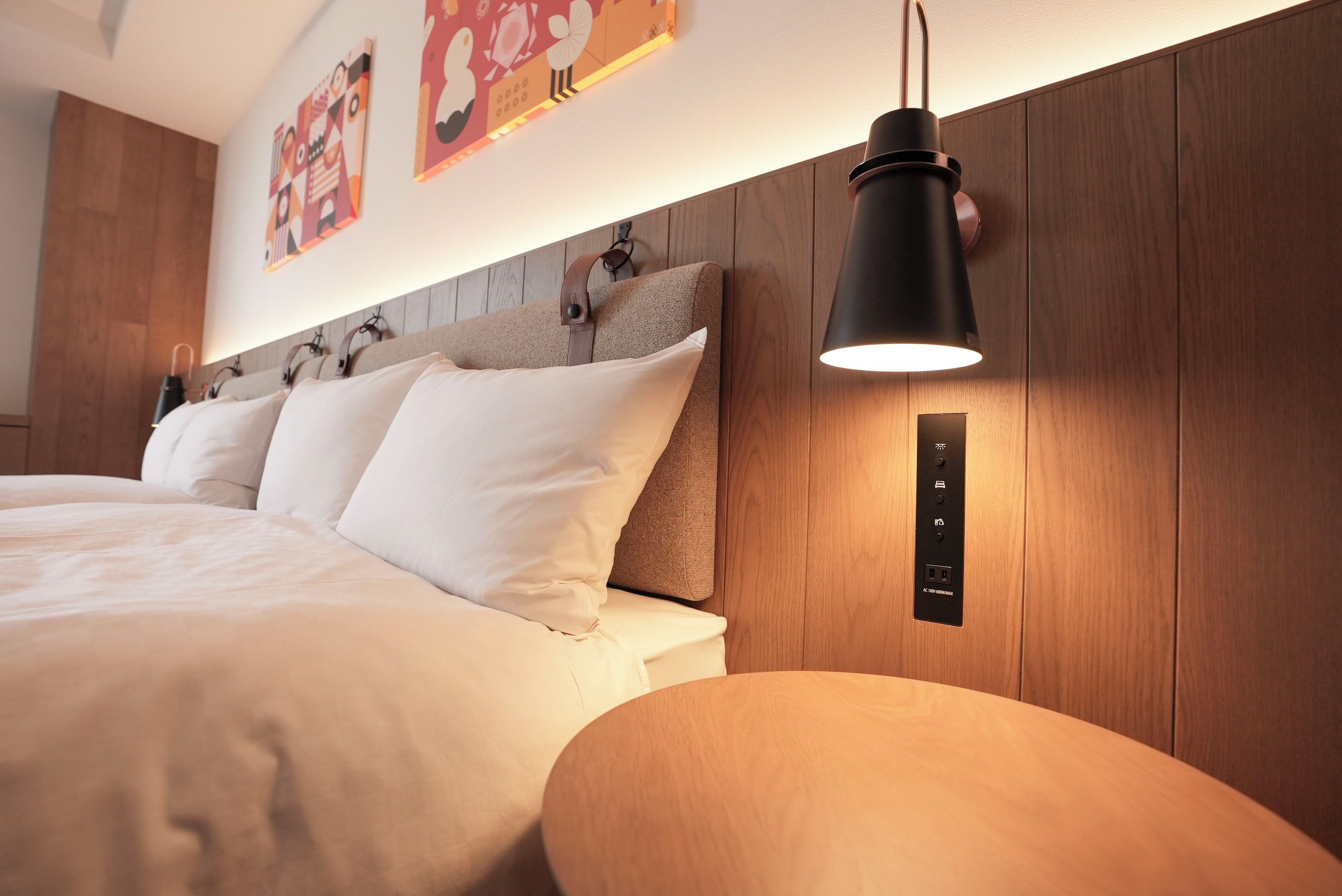A modern bedroom with a wooden headboard, a bed with white linens, a black pendant light, and a wall-mounted control panel.