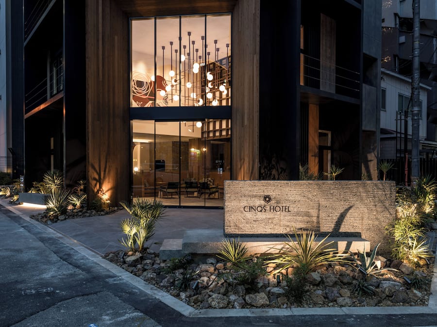 A modern hotel entrance with a glass facade, a wooden signboard, and a landscaped garden area.