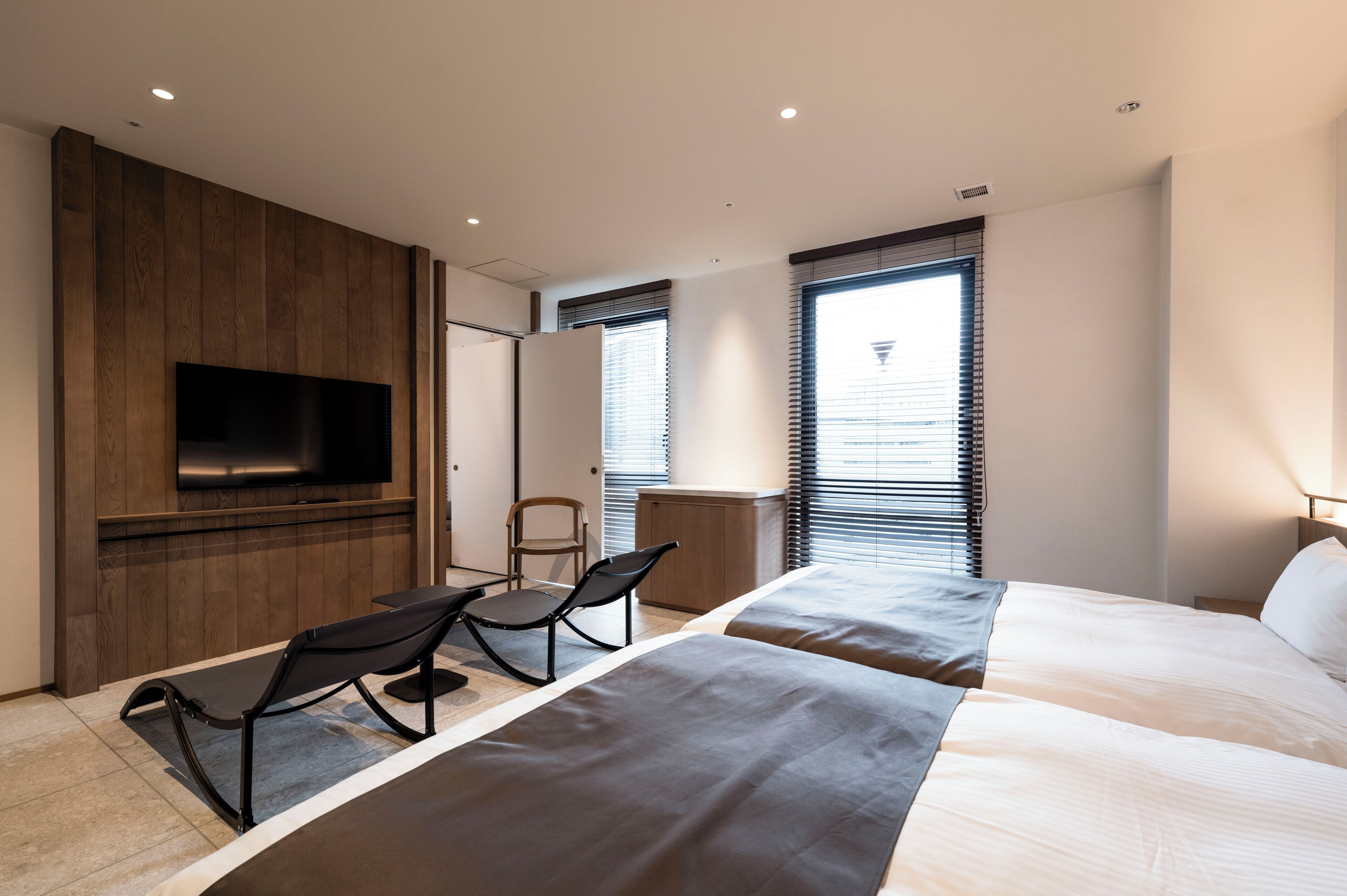 A modern hotel room with a large bed, a TV mounted on a wooden panel, a desk with a chair, and a window with blinds.