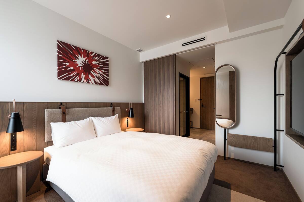 Quarto de hotel moderno com uma cama grande, uma mesa de cabeceira com abajur, um espelho fixado na parede e uma porta que dá para outro quarto.