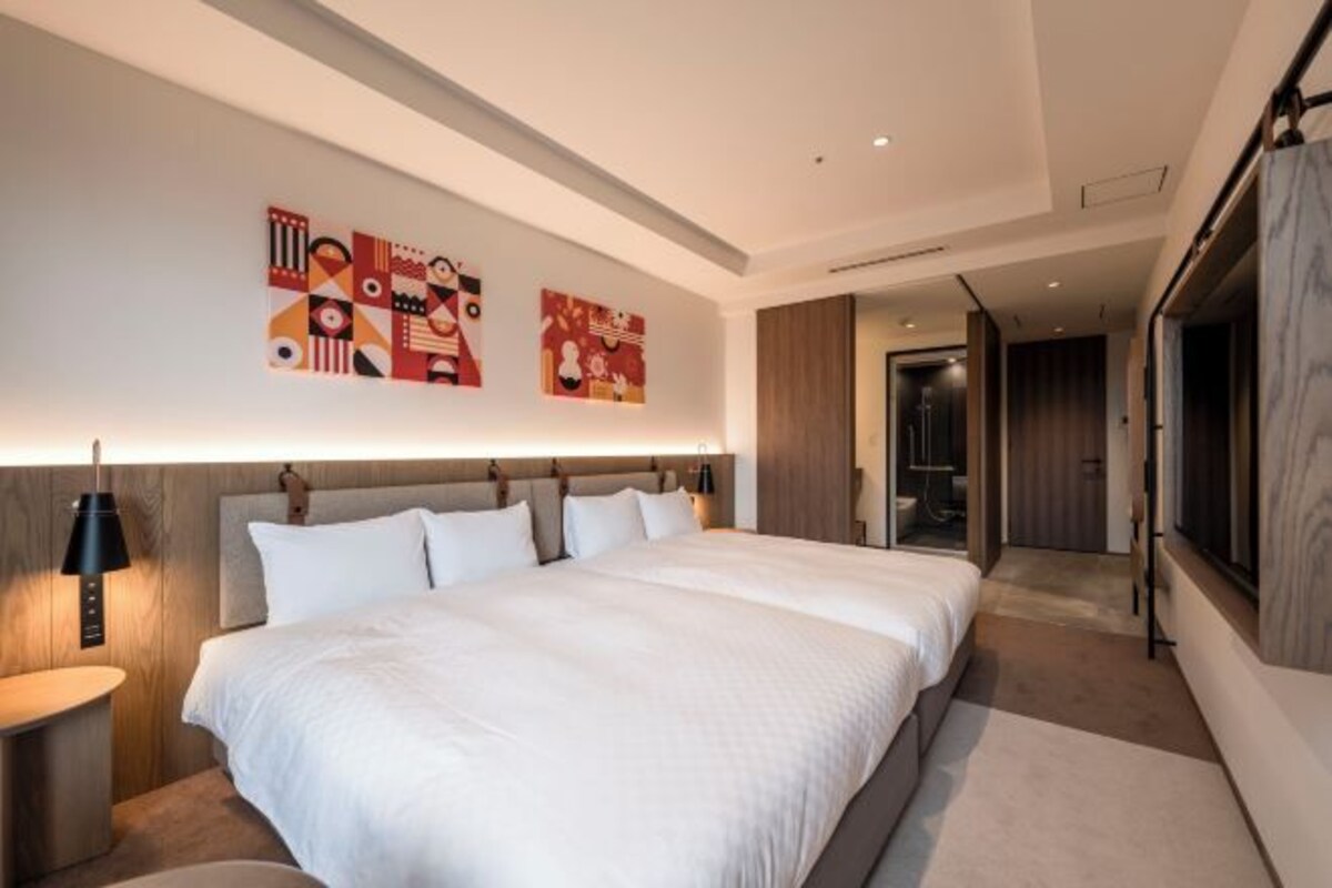 A modern hotel room with a large bed, a headboard with built-in lighting, a bedside table with a lamp, and abstract wall art.