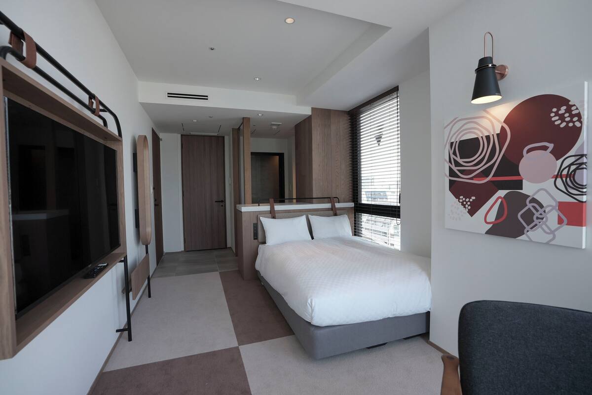 Quarto moderno com cama, televisão e uma janela grande com persianas.
