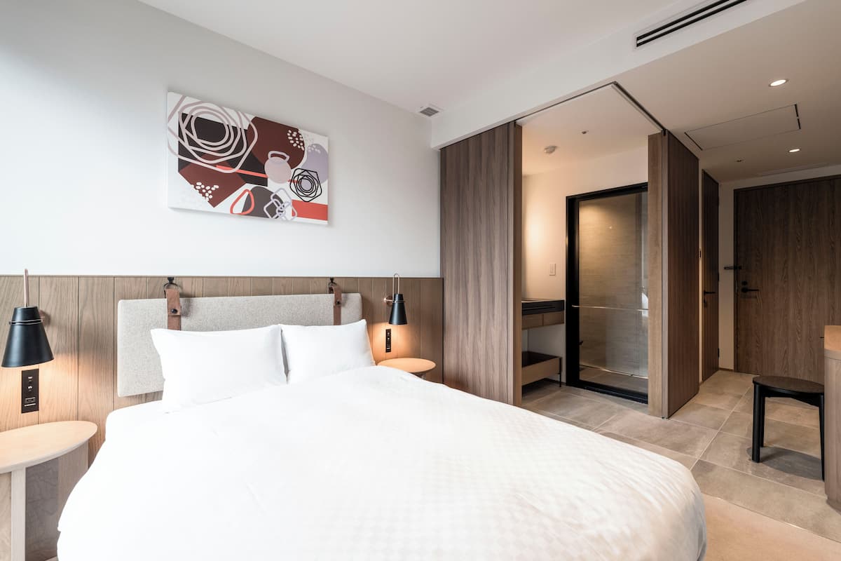 Quarto de hotel moderno com uma cama grande, cabeceira com detalhes em couro, uma obra de arte abstrata na parede, porta deslizante para um banheiro e uma mesinha de cabeceira com abajur.