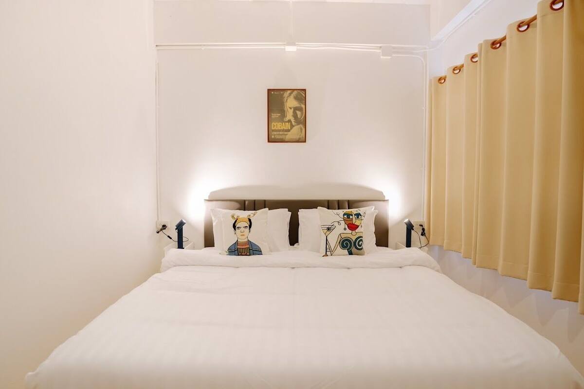 Una habitación de hotel con una cama, dos almohadas con personajes de caricatura, un cartel en la pared y cortinas.