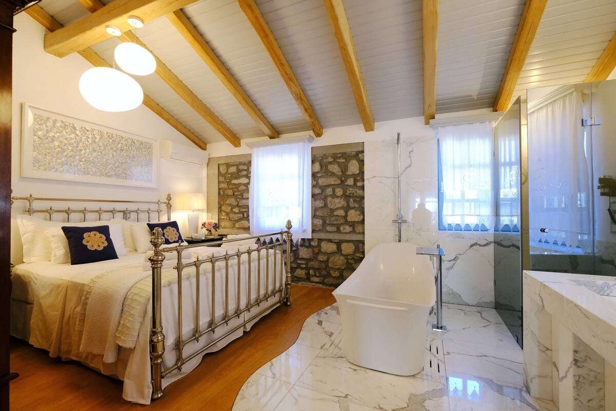 Un dormitorio con cama, un ventanal grande, pared de piedra, bañera y suelo de mármol.