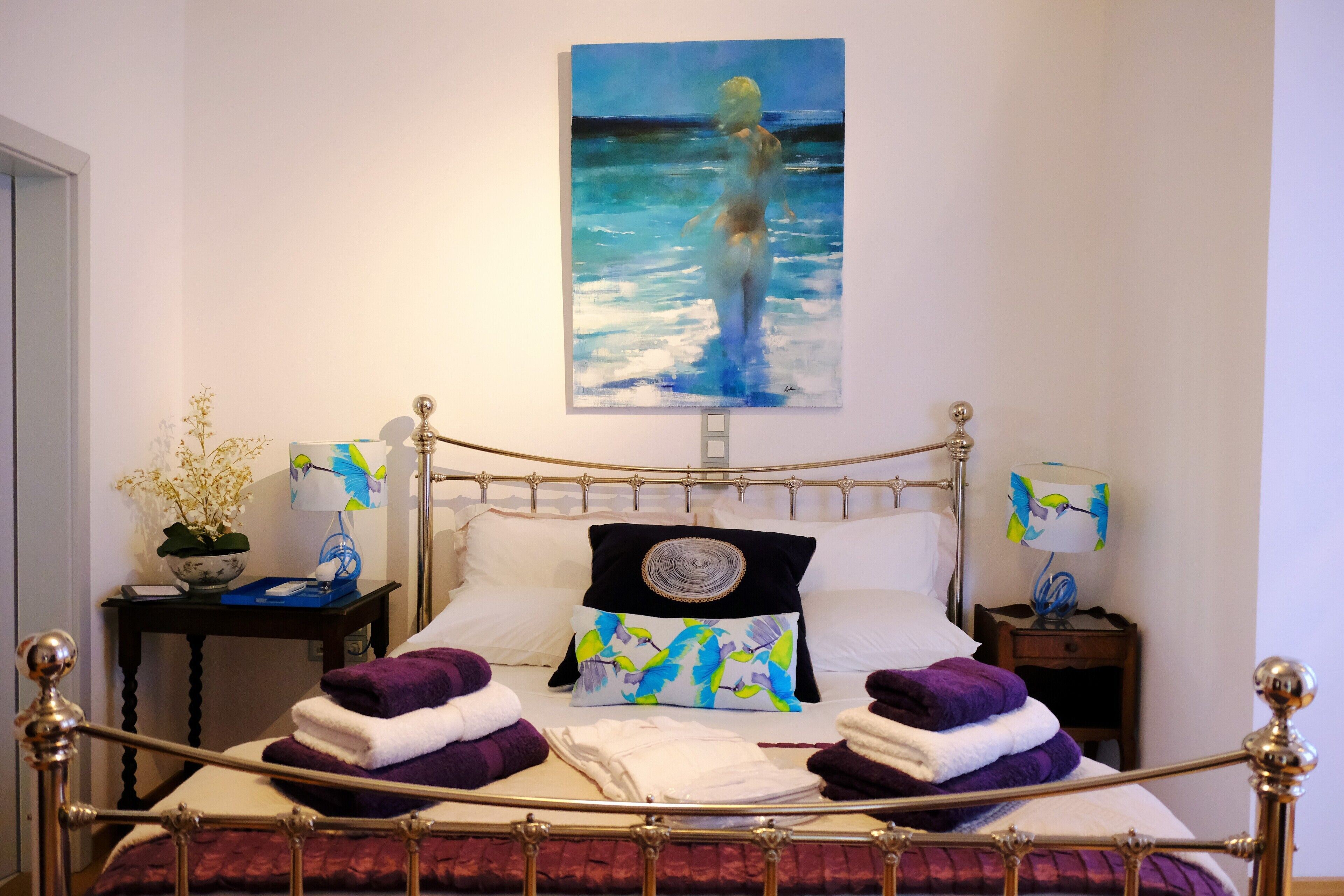A neatly made bed with a painting of a person in water, a lamp, and a small table with a plant.