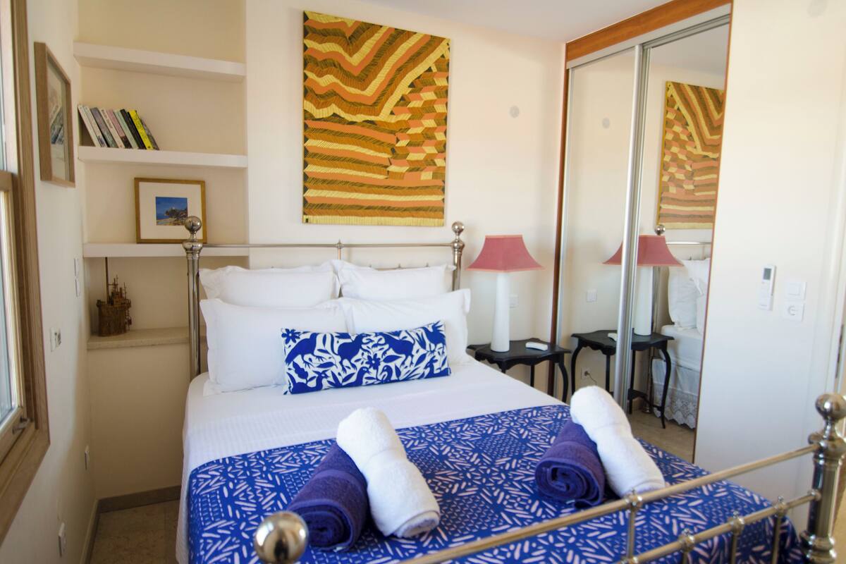 Una cama bien hecha con ropa de cama blanca y azul estampada, una mesita de noche con lámpara y una pared con un cuadro de motivos geométricos.