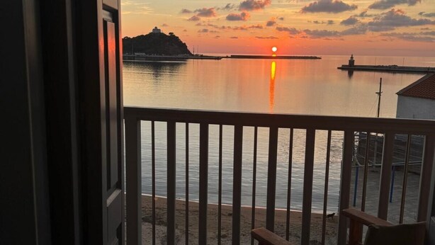 Varanda com vista para um pôr do sol sobre águas calmas, uma cadeira de madeira e uma ilha distante com um farol.