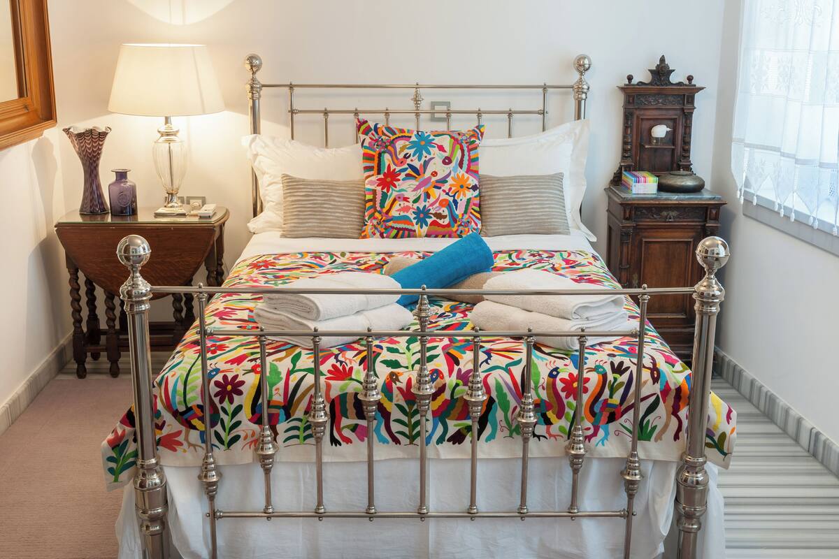 Una cama bien hecha con un colorido edredón floral, un cabecero metálico y una mesita de noche con una lámpara y objetos decorativos.