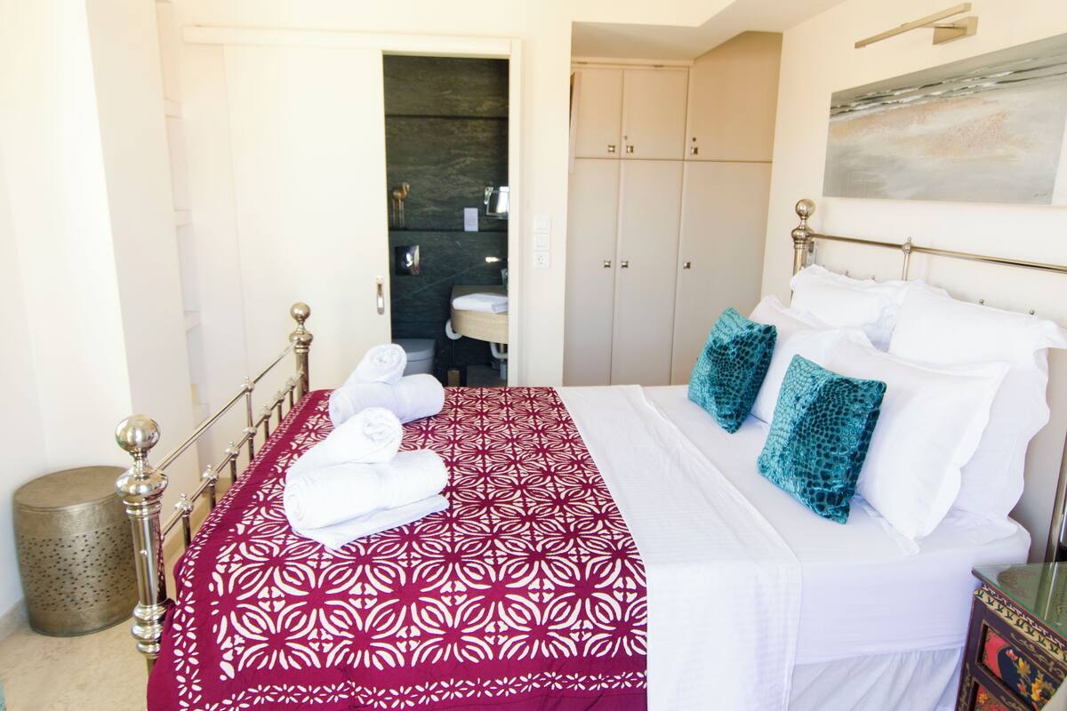 Una cama bien hecha con una colcha estampada, un baño visible a través de una puerta abierta y un cuadro en la pared.