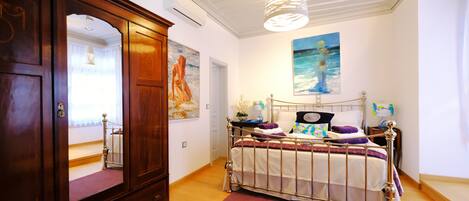 A bedroom with a wooden wardrobe, a metal bed frame, a large mirror, and a painting of a woman on the wall.