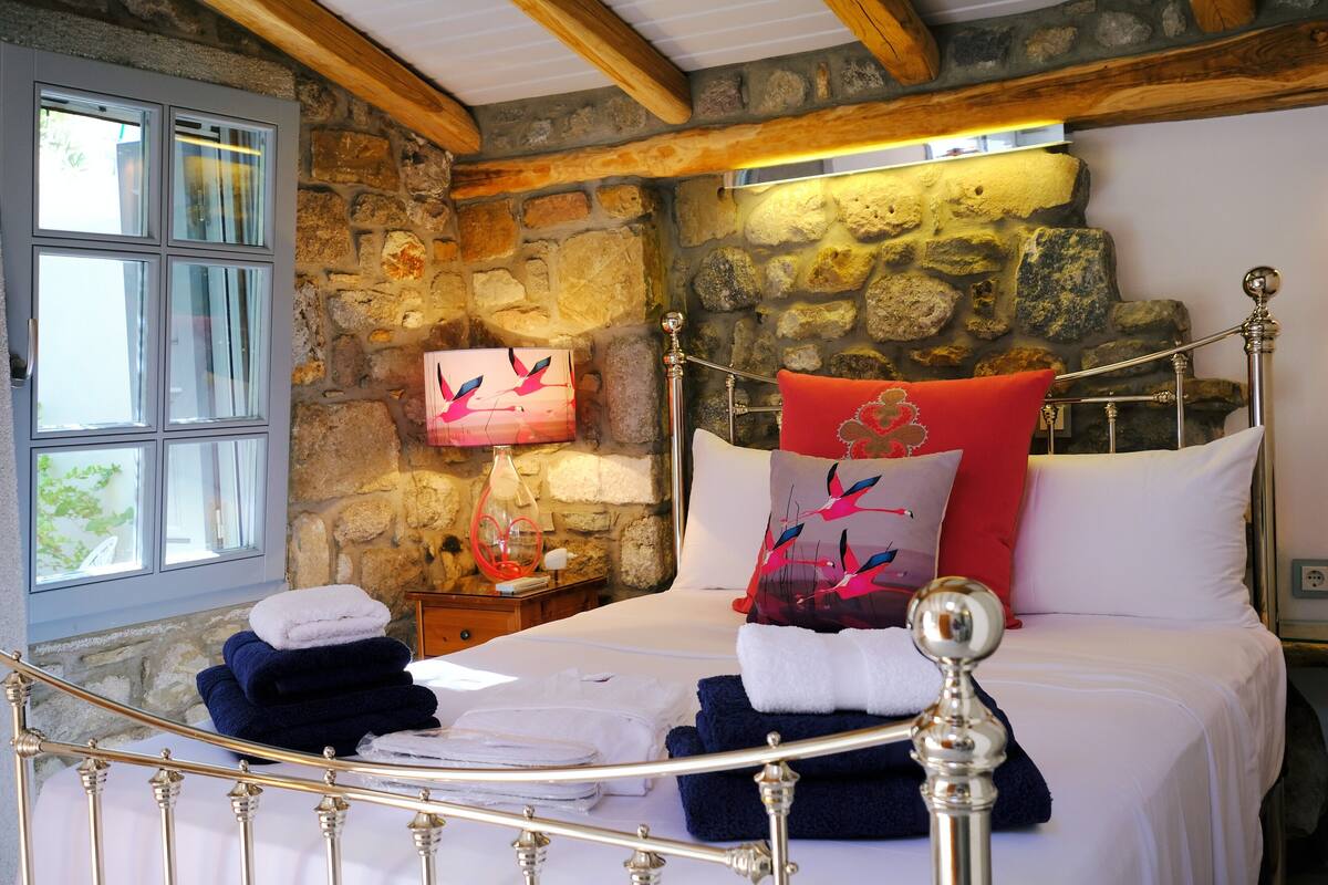 Un dormitorio con paredes de piedra, una cama con ropa de cama blanca, cabecero de metal y una mesita de noche con lámpara y objetos decorativos.