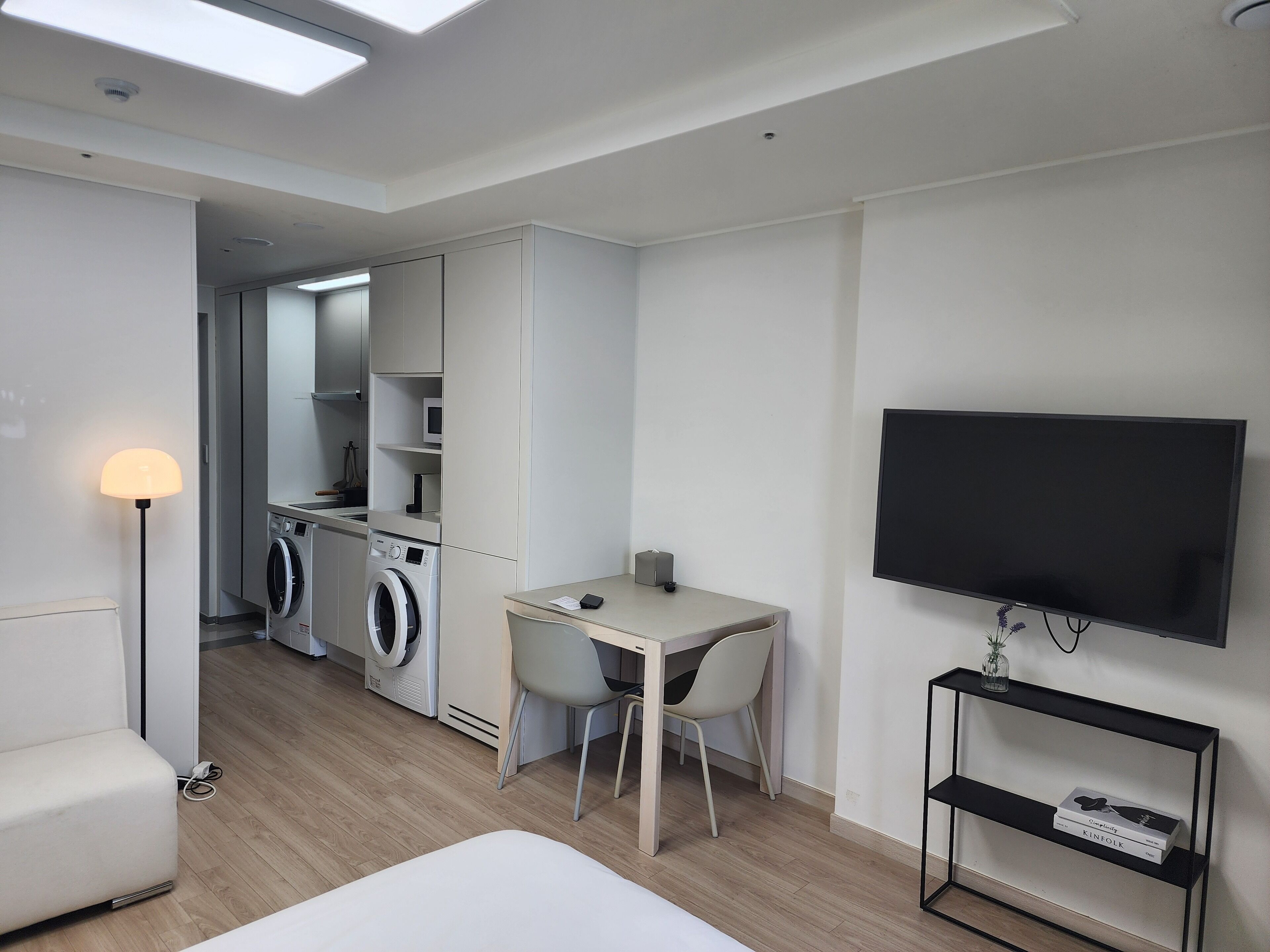 Une chambre d’hôtel moderne équipée d’un lit, d’un bureau avec des chaises, d’une télévision, d’un lave-linge et d’une étagère remplie de livres.