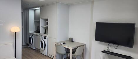 Une chambre d’hôtel moderne équipée d’un lit, d’un bureau avec des chaises, d’une télévision, d’un lave-linge et d’une étagère remplie de livres.