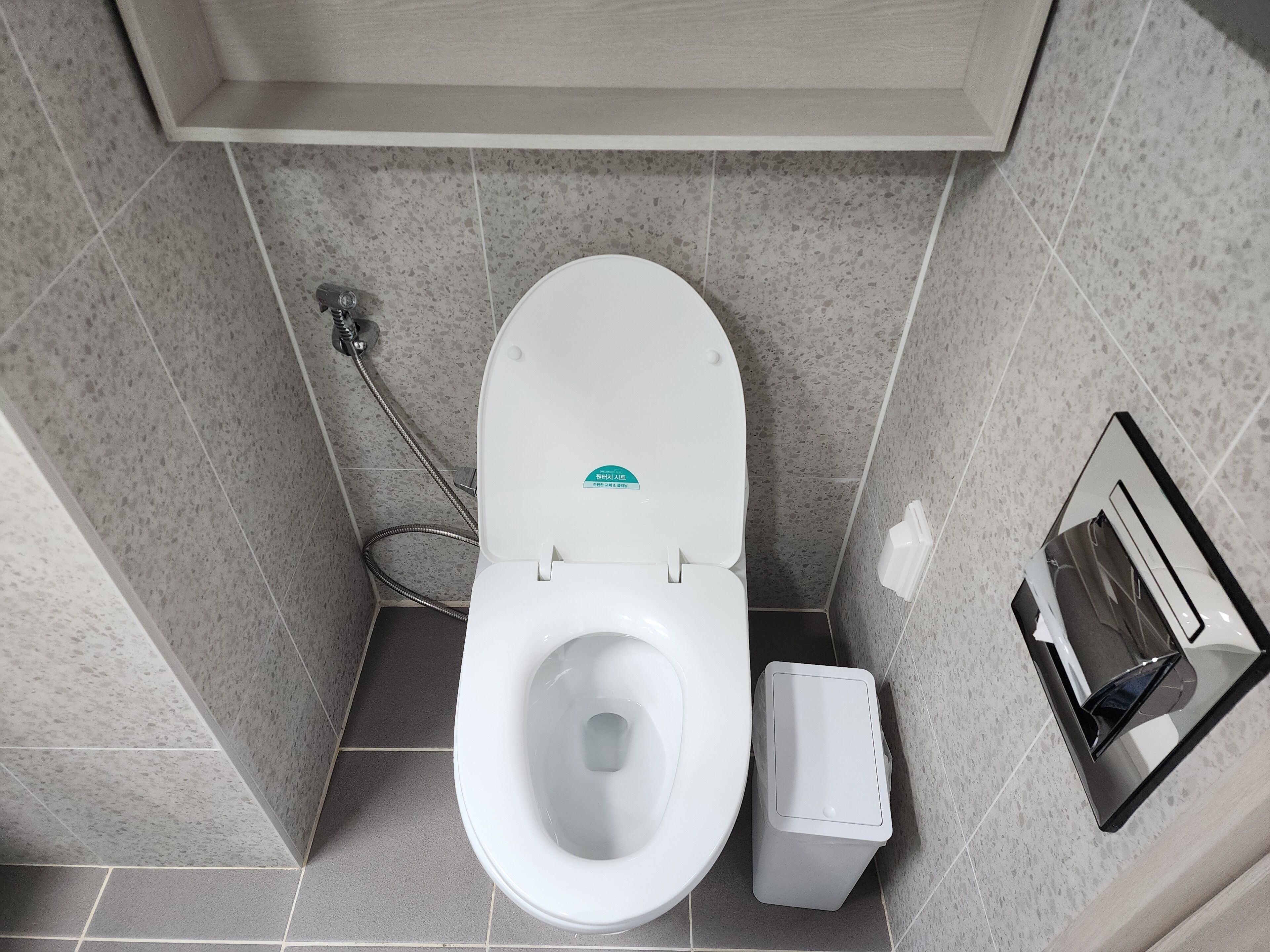 A modern bathroom with a white toilet, a shower head, and a trash bin.