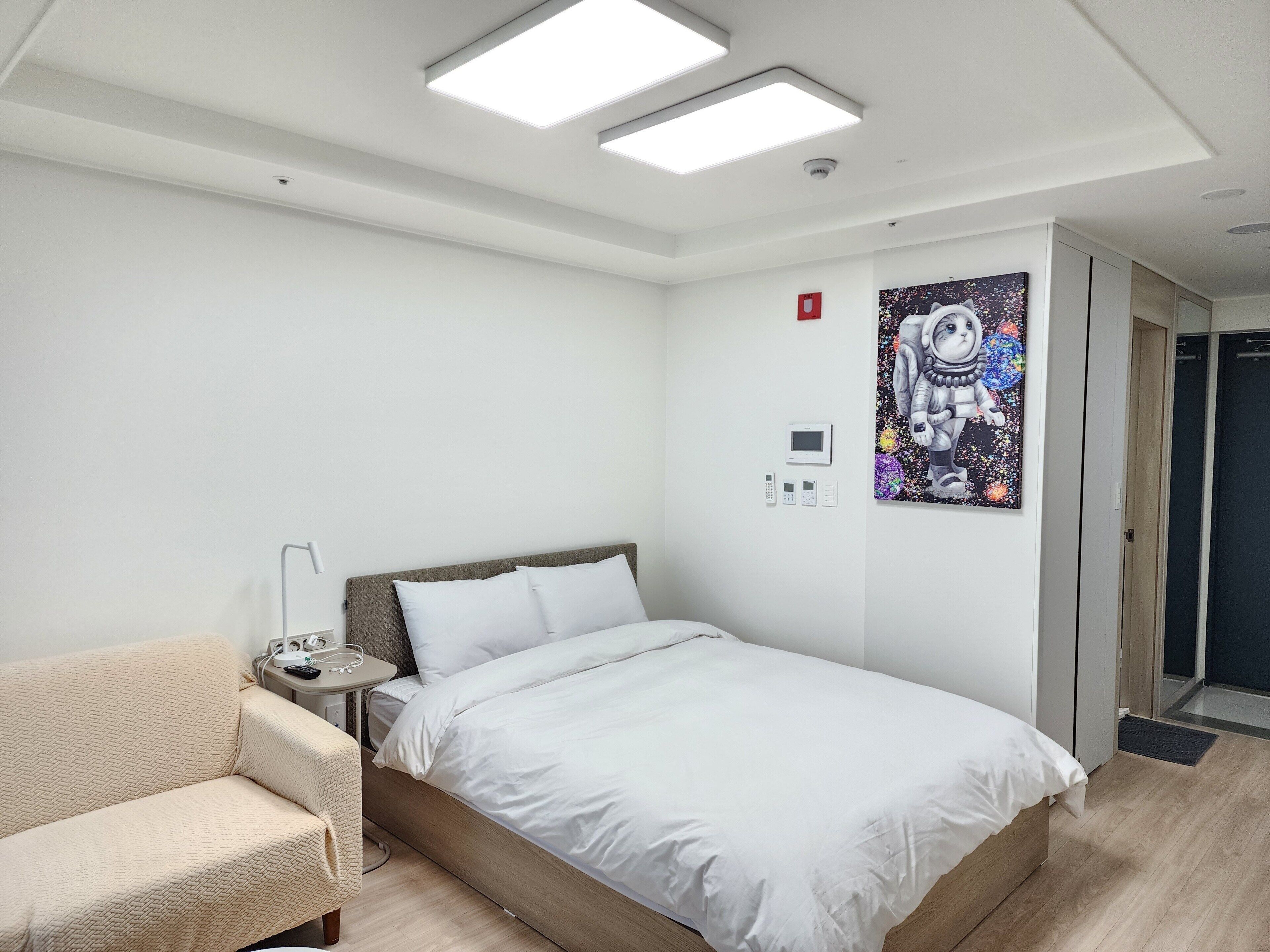 Moderni hotellihuone, jossa on sänky, yöpöytä, tuoli, taulu seinällä ja ovi oikealla puolella.