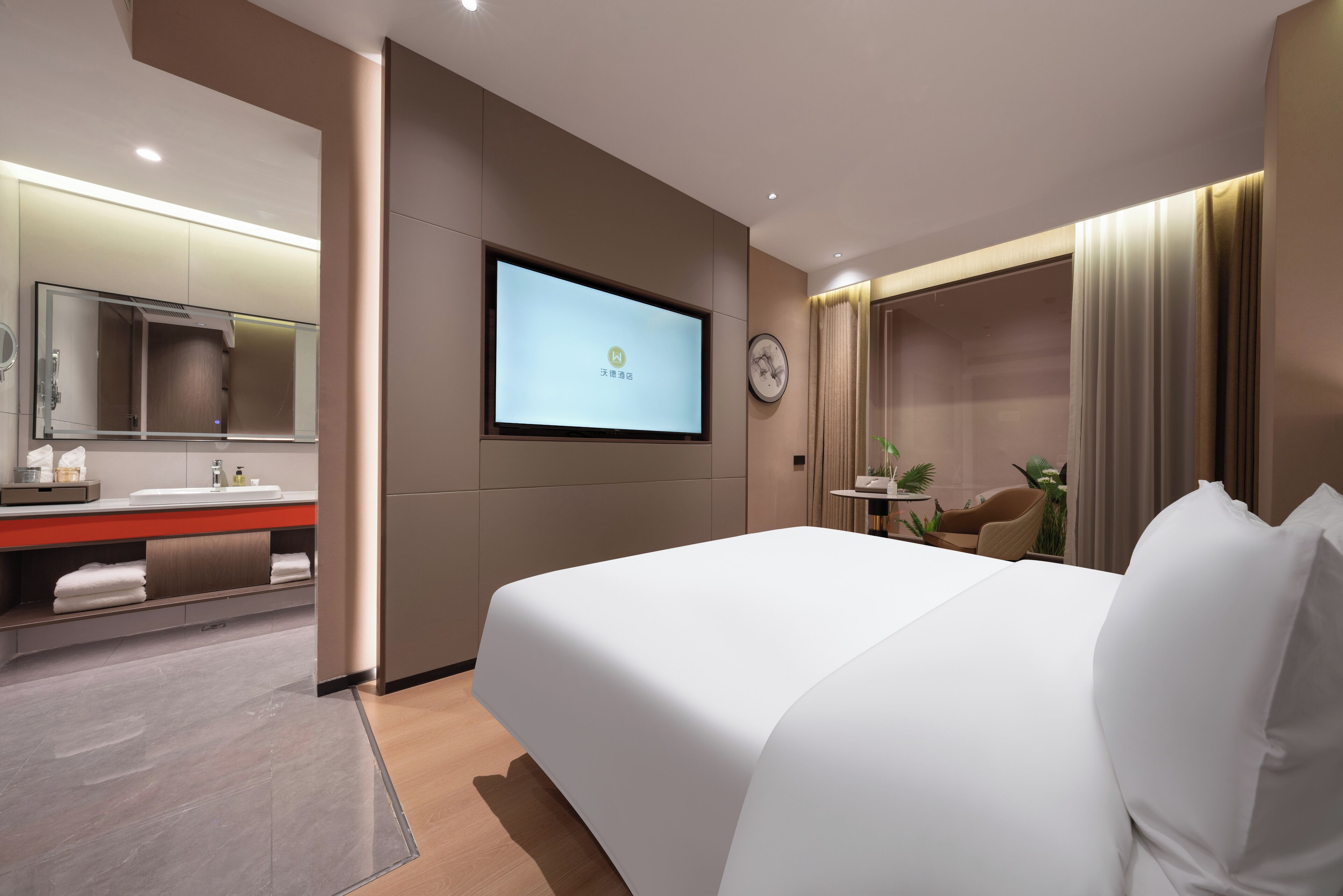 Een moderne hotelkamer met een groot bed, een aan de muur bevestigde televisie, een badkamer met spiegel en wastafel, en een klein tafeltje met een plant.