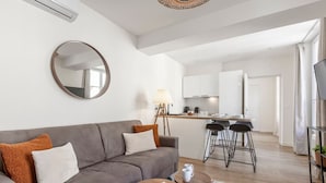 Ein modernes Wohnzimmer mit einem grauen Sofa, einem runden Spiegel und einem kleinen Couchtisch. Im Hintergrund ist ein Küchenbereich mit weißen Schränken und einem Esstisch zu sehen.