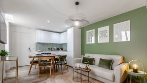 A modern living room with a white sofa, a dining area with a wooden table and chairs, a kitchen with white cabinets, and a green accent wall with framed artwork.