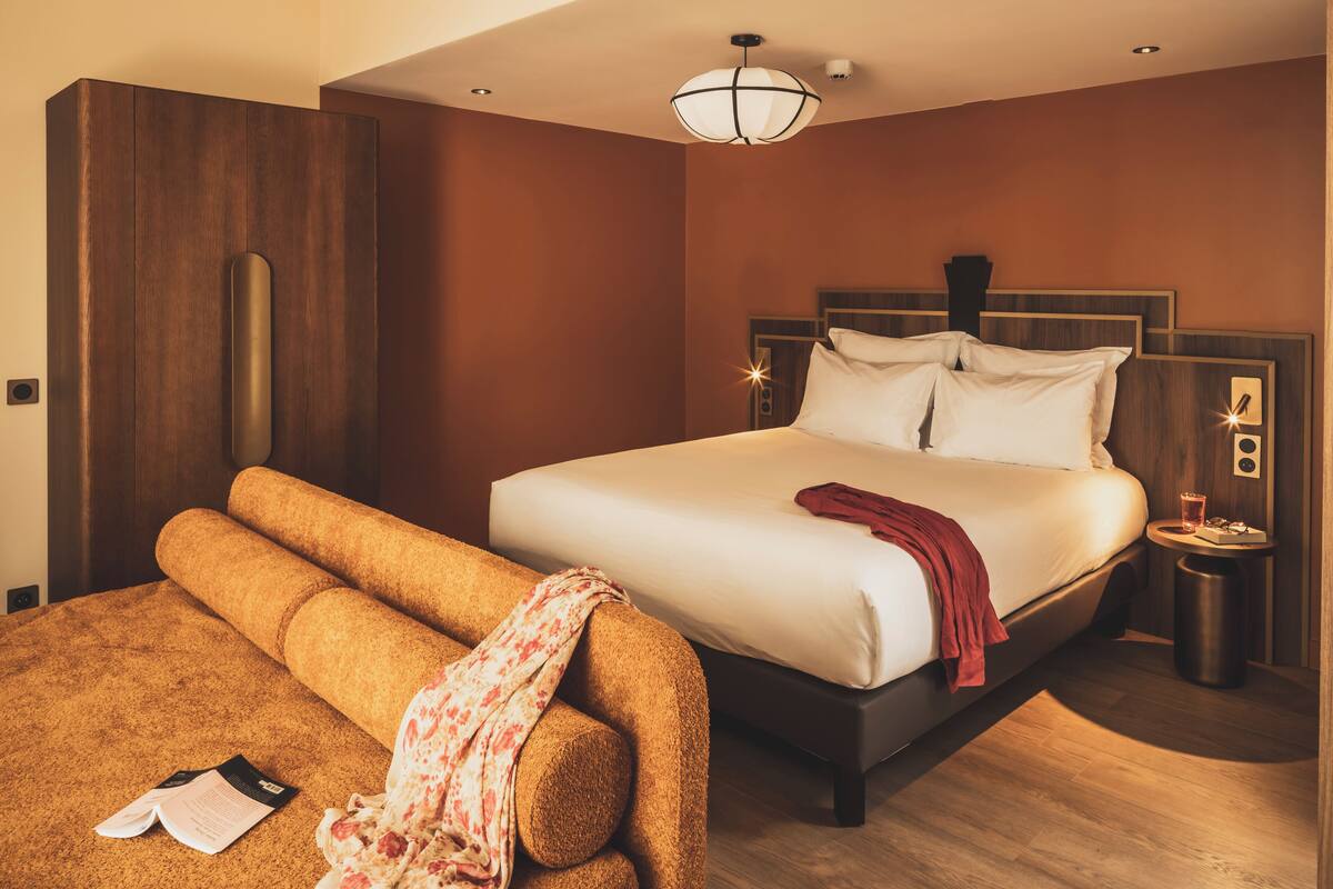 A hotel room with a large bed, a wooden wardrobe, a small bedside table with a lamp, and a reading area with a sofa and a book.