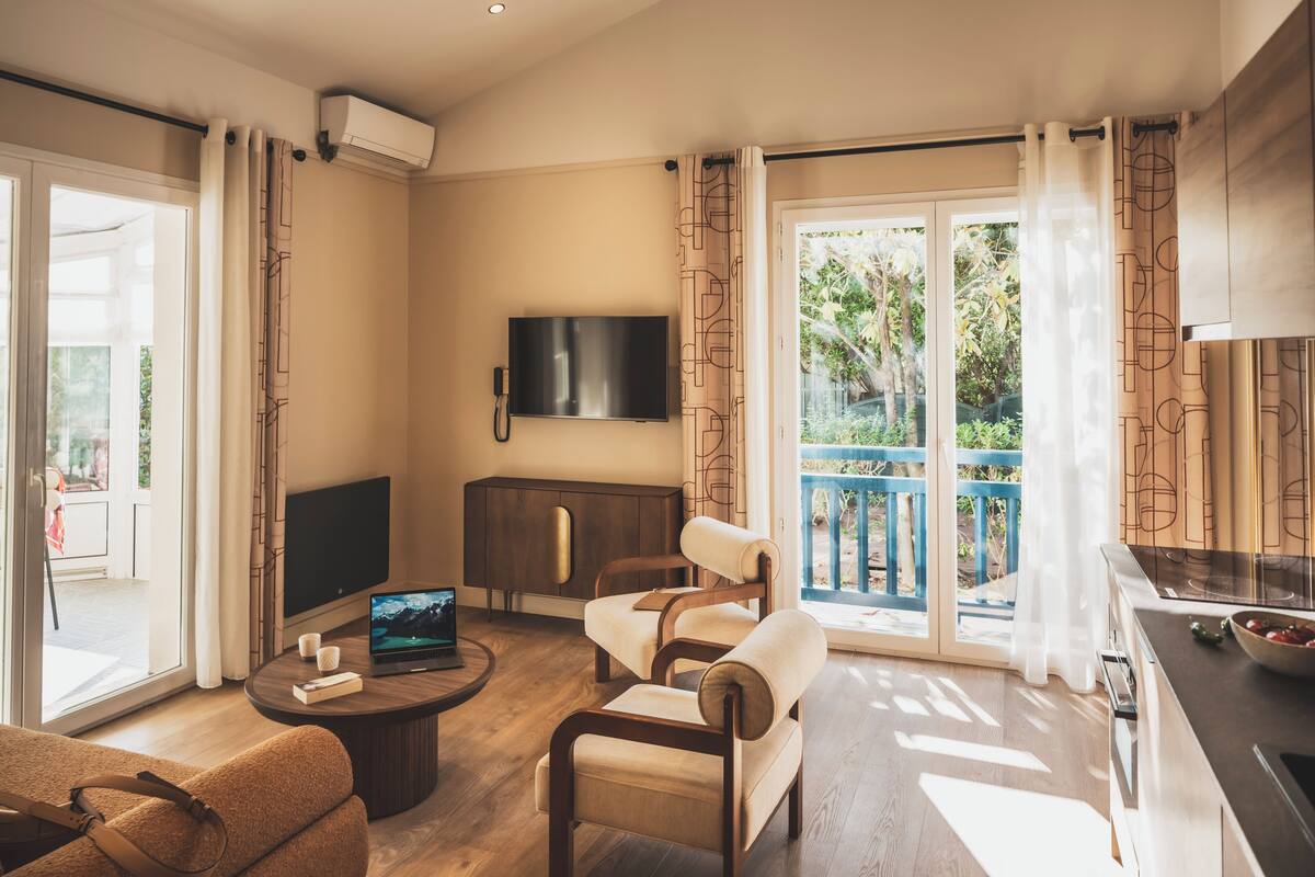A modern living room with a flat-screen TV, a sofa, a coffee table, and a balcony with a view of greenery.