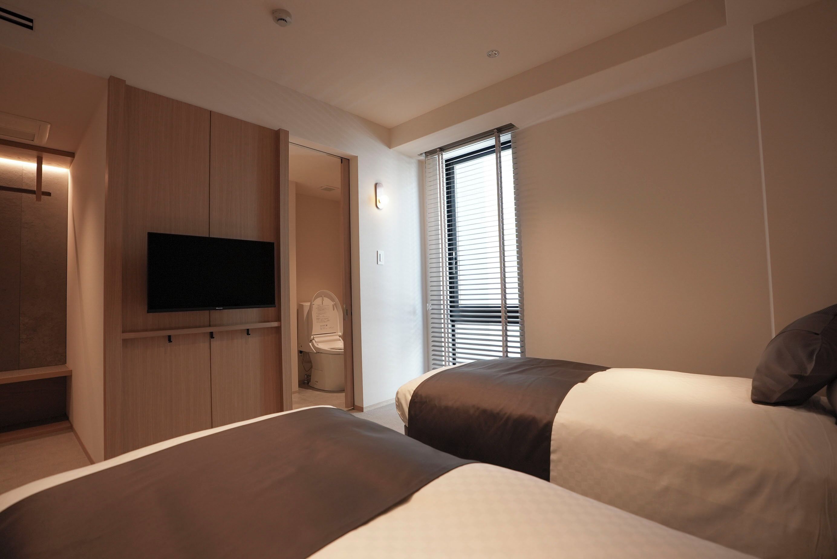 A hotel room with two beds, a flat-screen TV mounted on the wall, and a window with blinds.