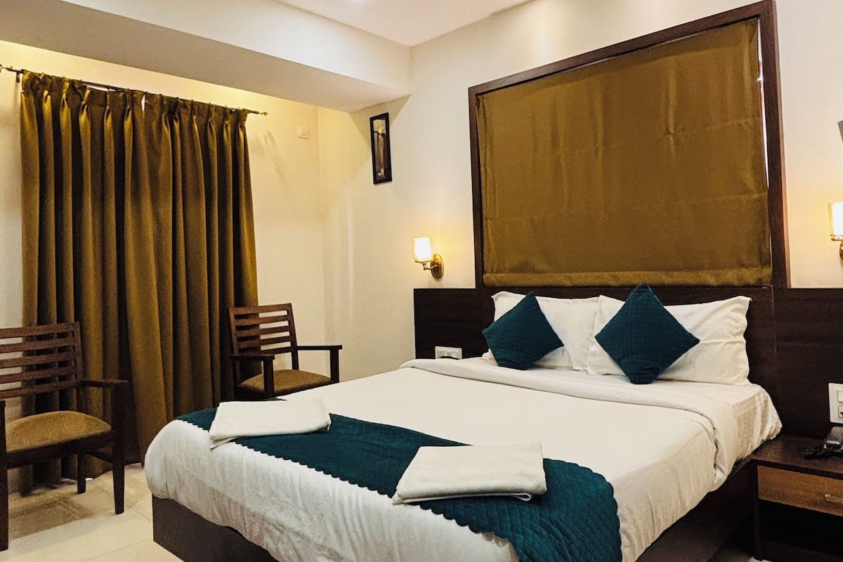 A hotel room with a bed, a ceiling fan, a nightstand, a chair, a desk, and a window with curtains.