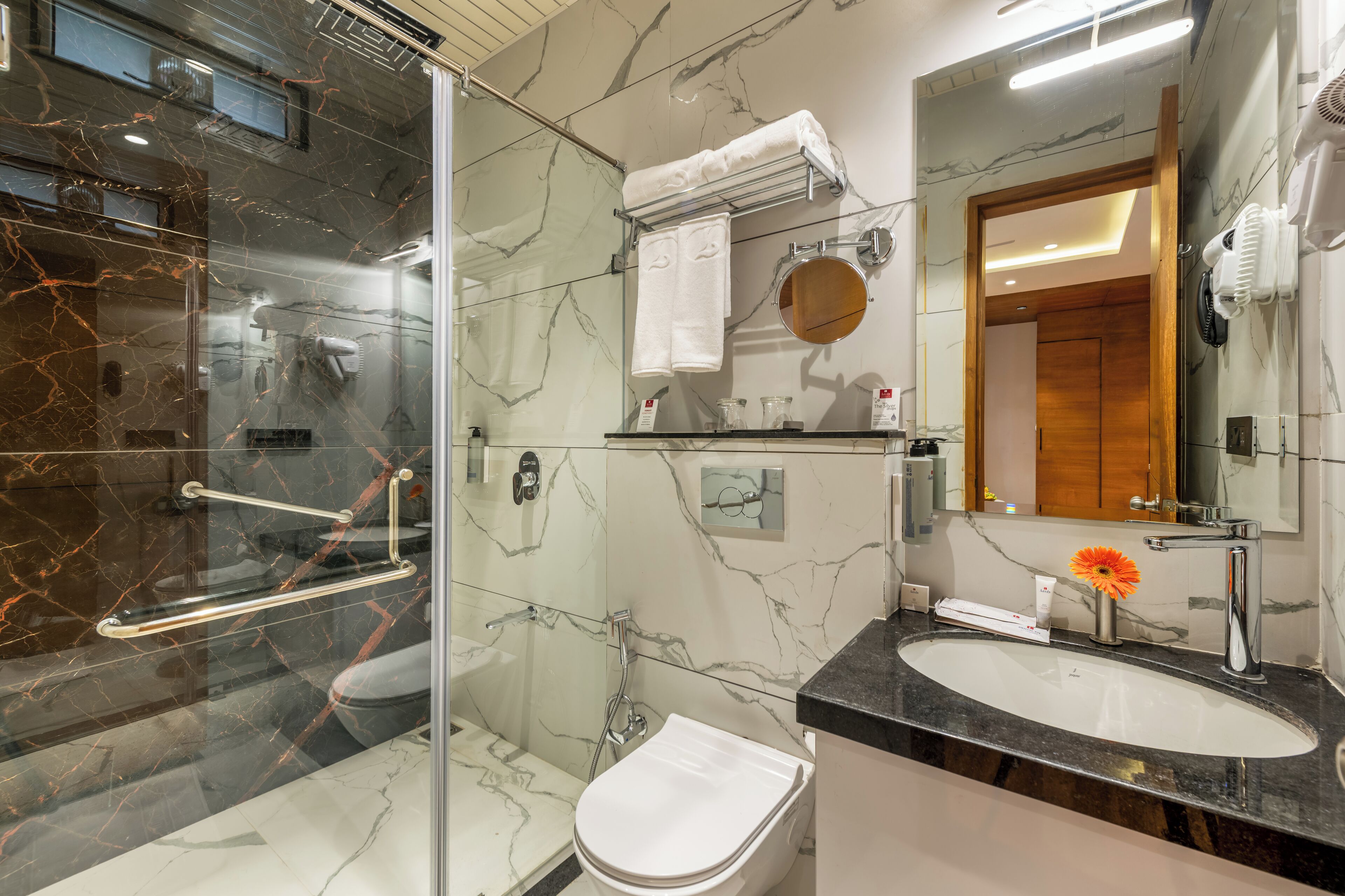 Un baño moderno con cabina de ducha de vidrio, inodoro blanco, pared con mosaicos de mármol, lavabo con encimera negra y espejo.