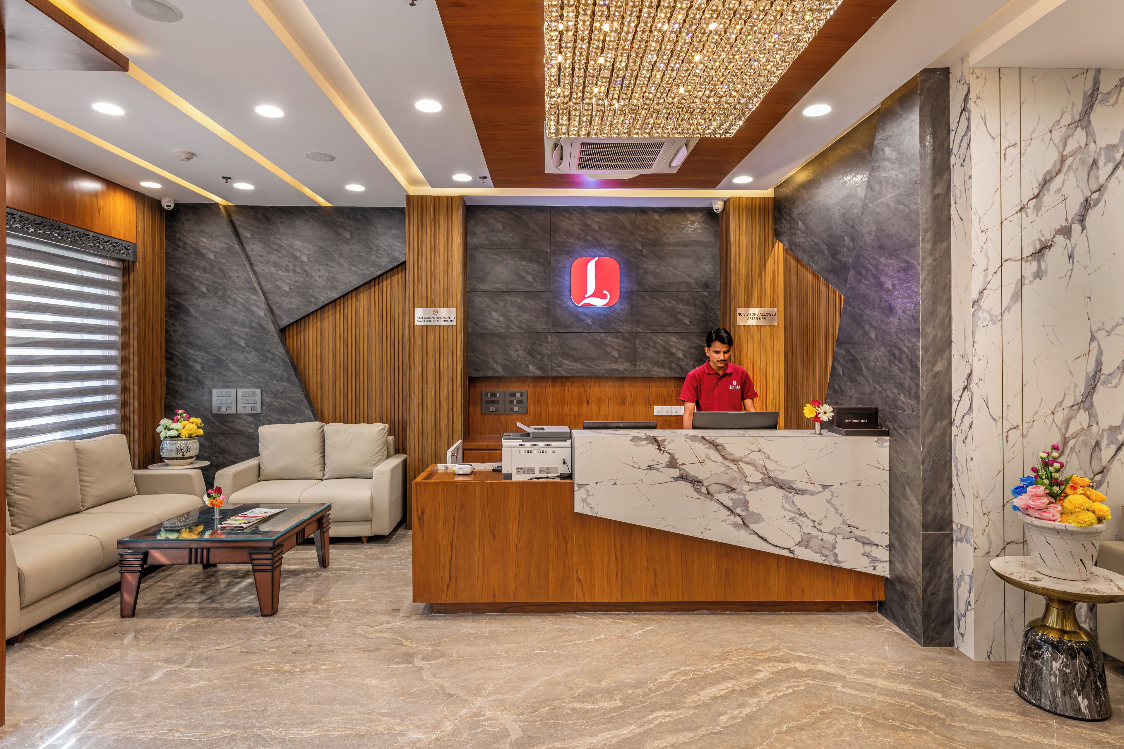A hotel reception area with a wooden reception desk, a receptionist, a sofa, a coffee table, and a decorative vase with flowers.