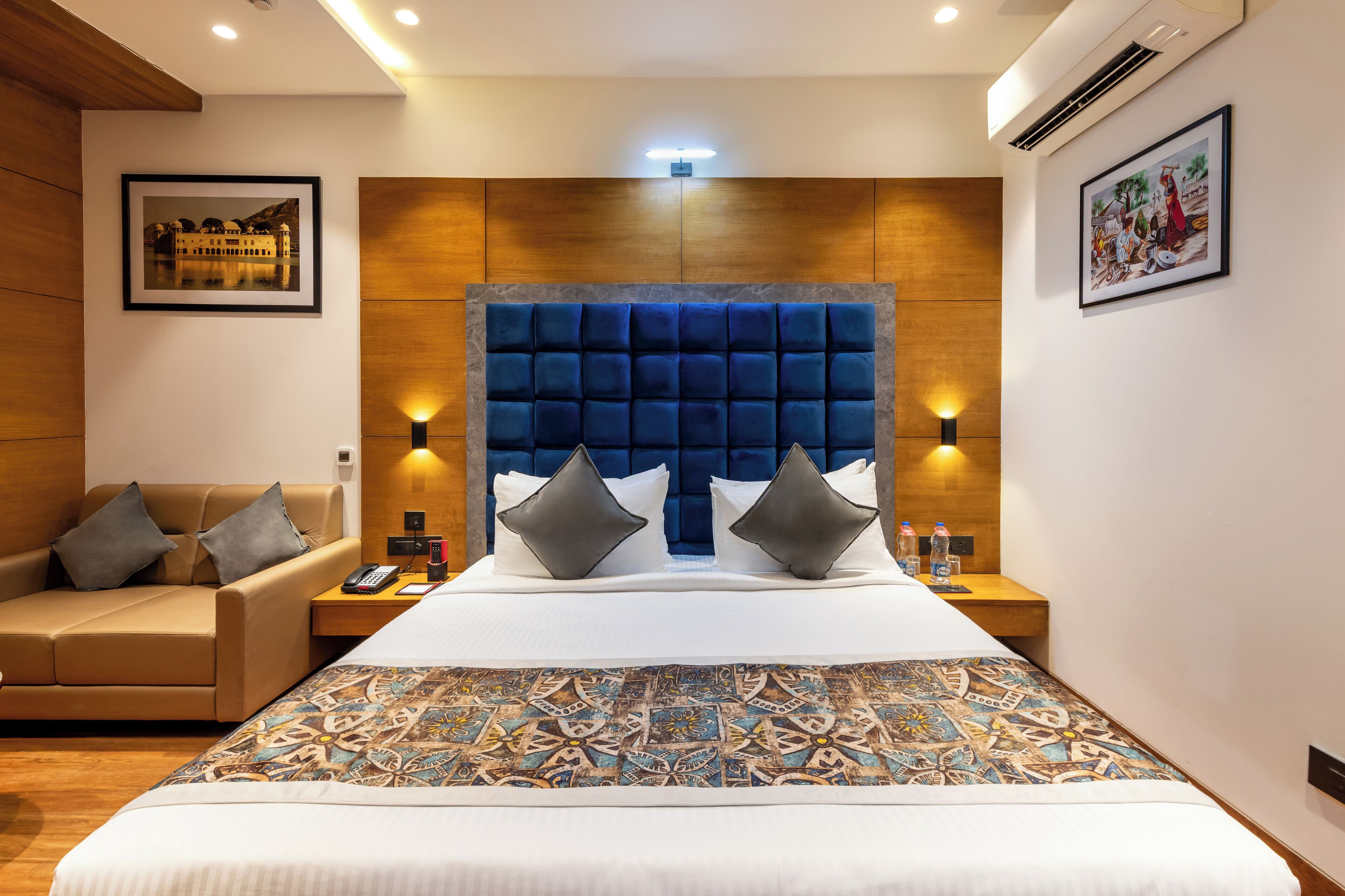 Habitación de hotel con una cama grande, un sofá, una mesita de noche con un teléfono y botellas de agua, y cuadros en las paredes.