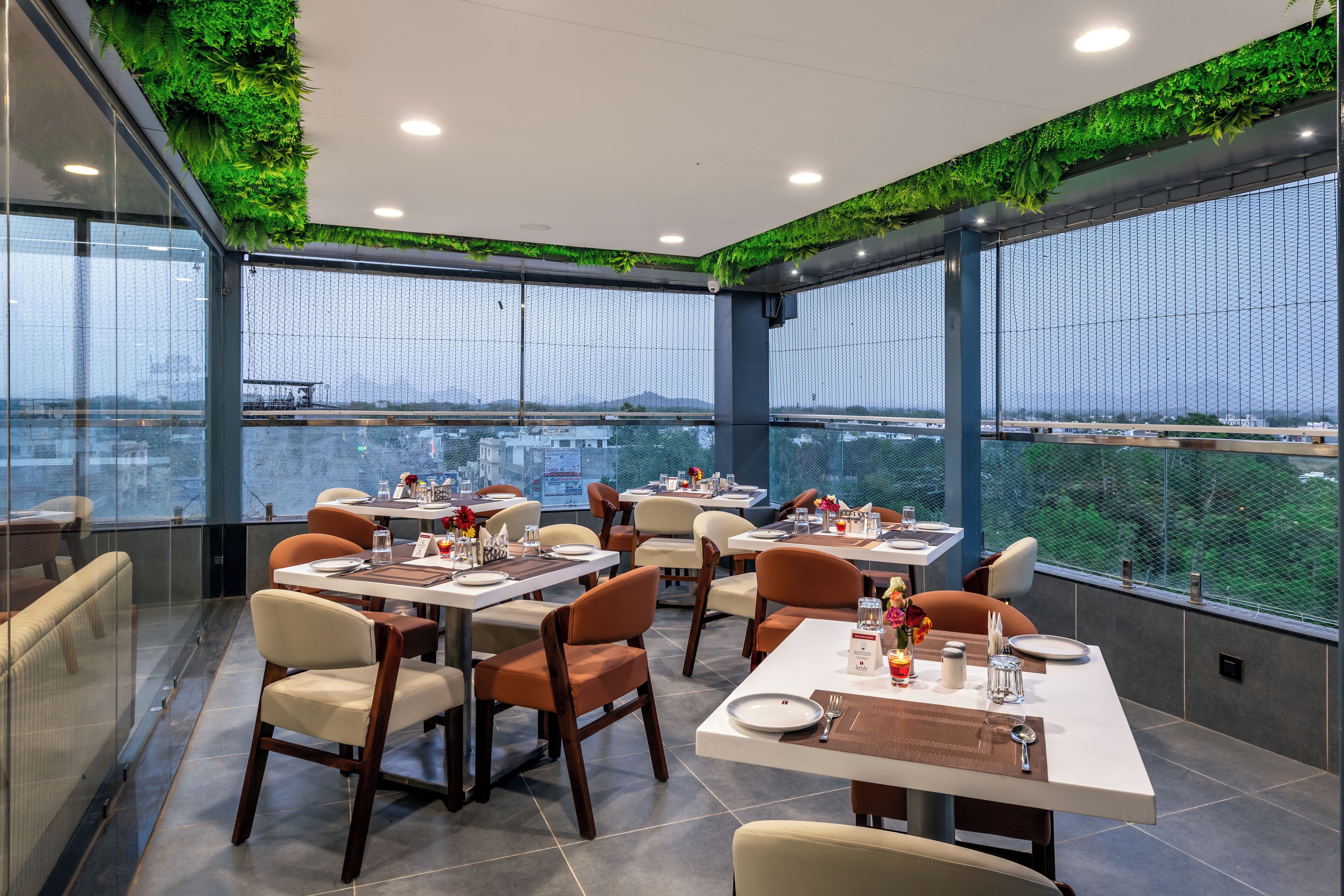 Un restaurante moderno con mesas y sillas, ventanas amplias y plantas verdes en el techo.