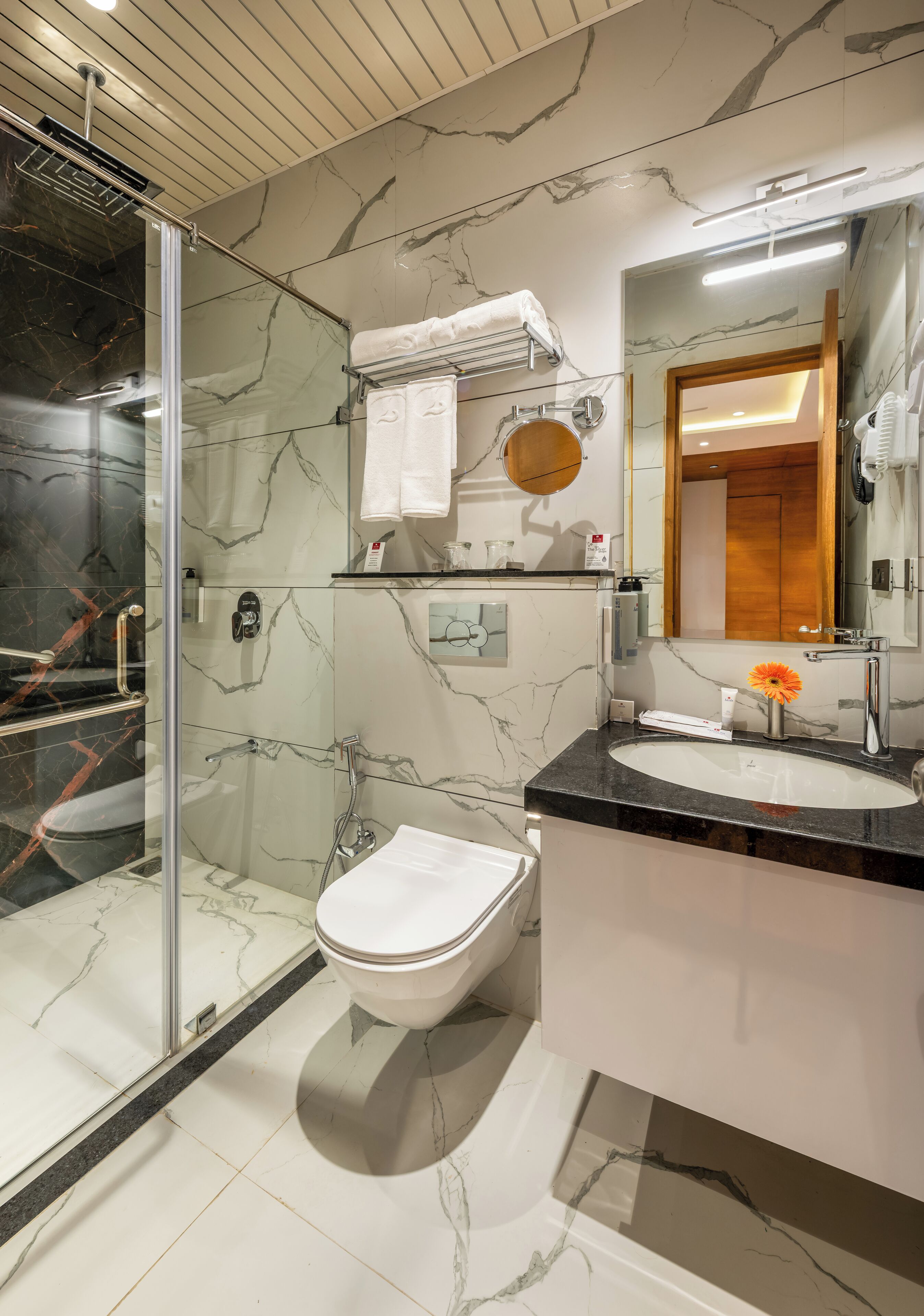 Un baño moderno con ducha envuelta en vidrio, piso de mármol, inodoro blanco y lavamanos con espejo.