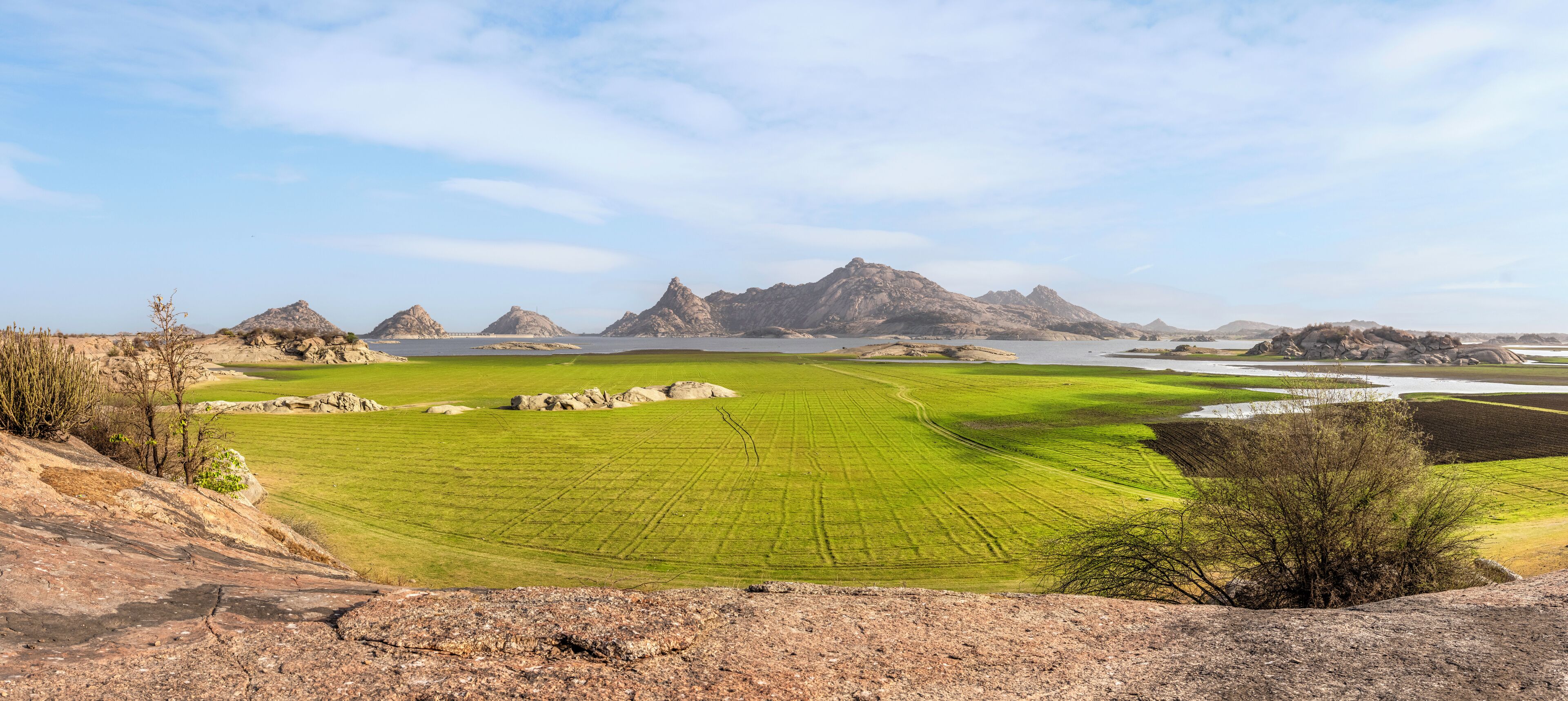 A panoramic view of a coastal landscape with green fields, rocky outcrops, and distant mountains under a clear sky.