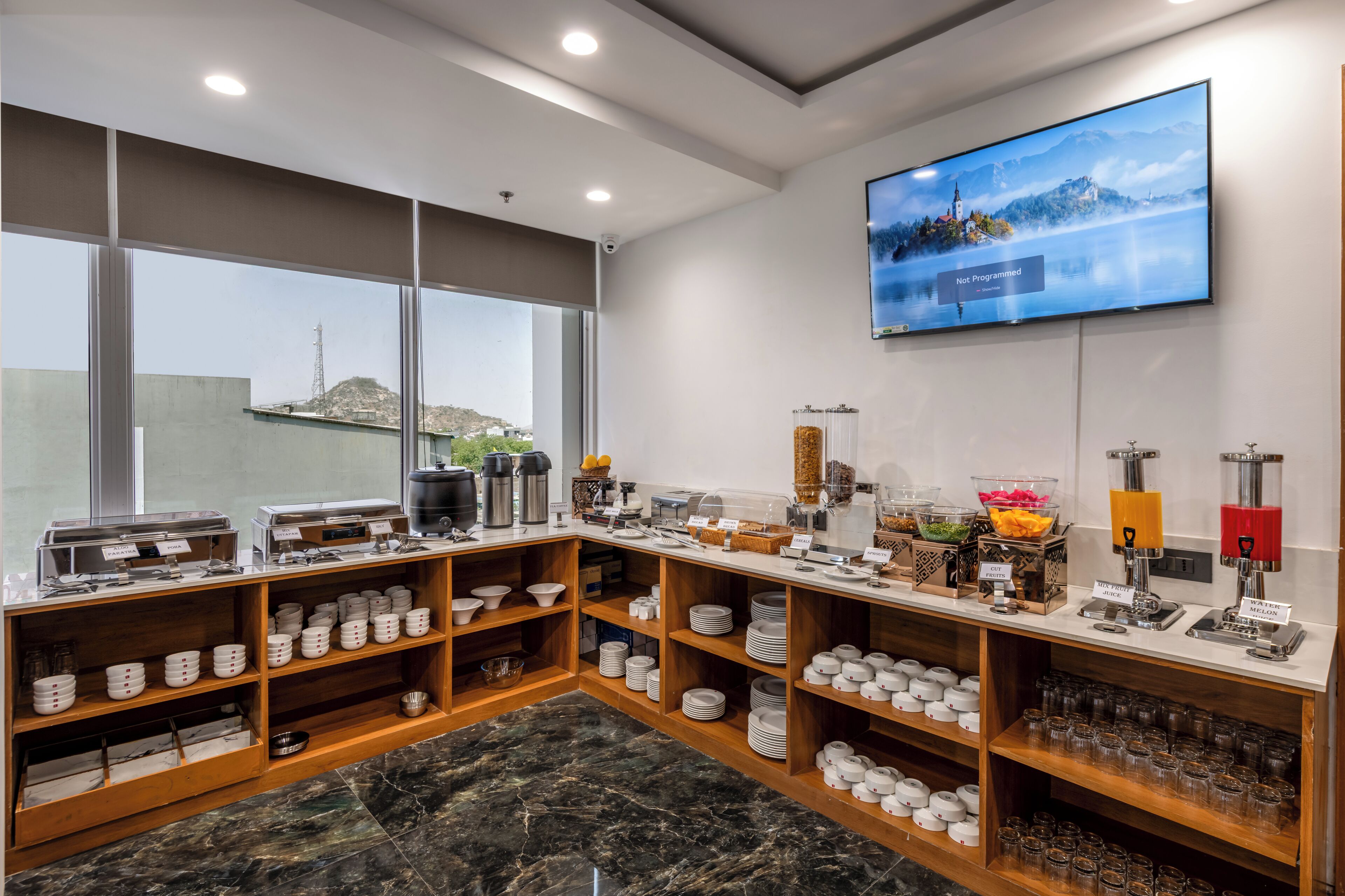 Zona de desayuno tipo buffet con diversas estaciones de comida, incluyendo dispensadores de café y zumo, y una gran pantalla de televisión mostrando un paisaje pintoresco.