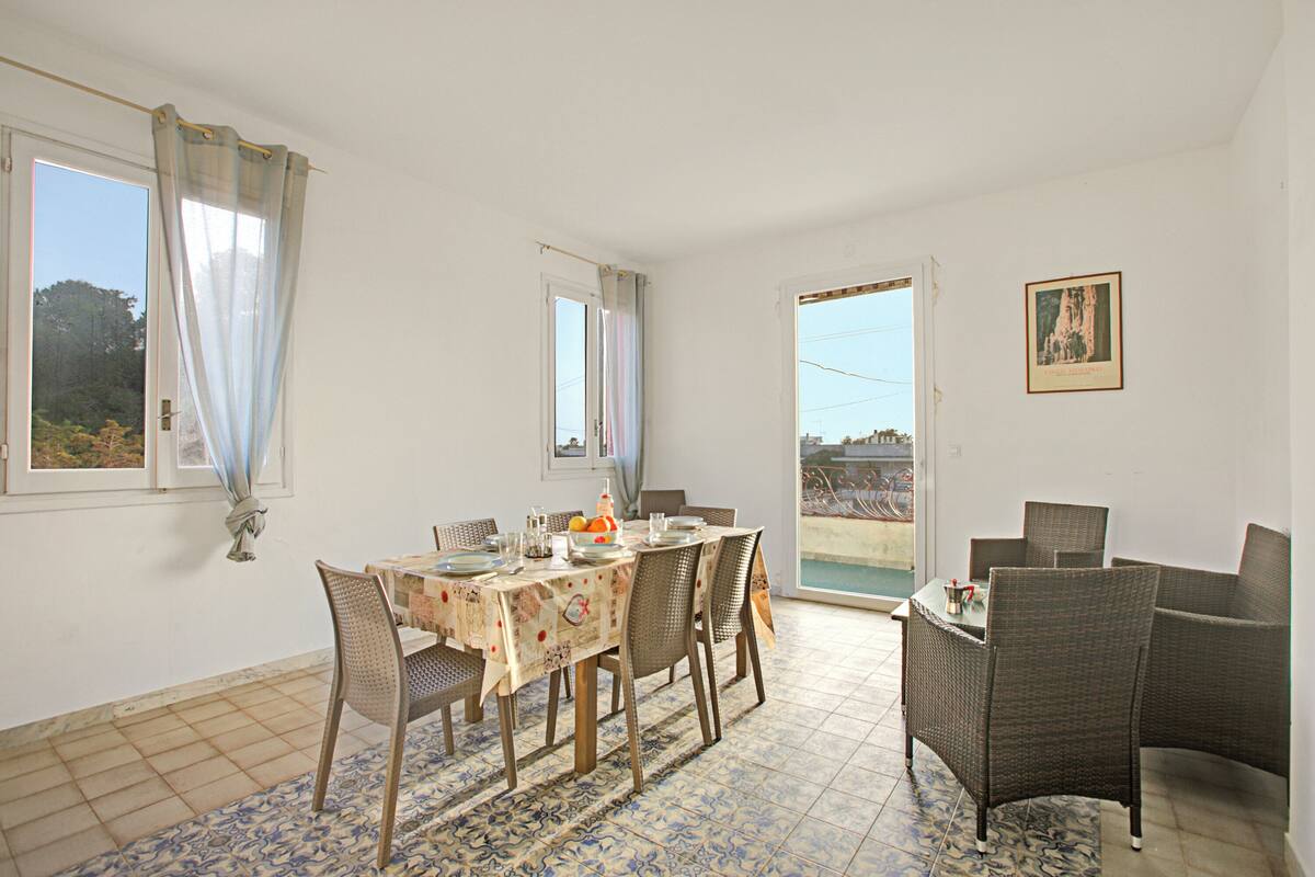 Un coin repas avec une table dressée pour quatre personnes, entouré de chaises en osier, d’un tapis à motifs et offrant une vue sur l’extérieur par une porte.