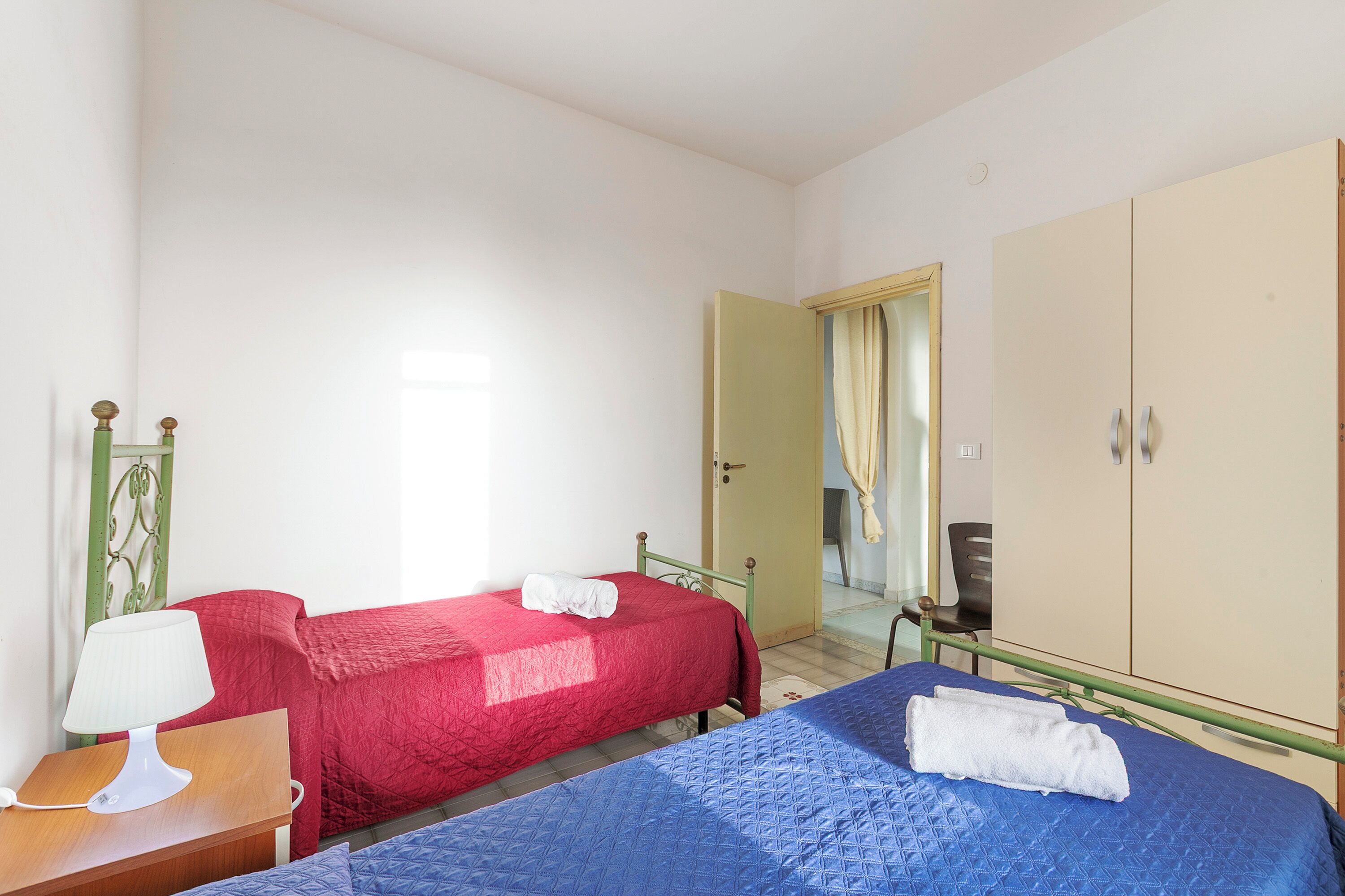 A room with two beds, one with a red bedspread and the other with a blue bedspread, a bedside table with a lamp, and a wardrobe.
