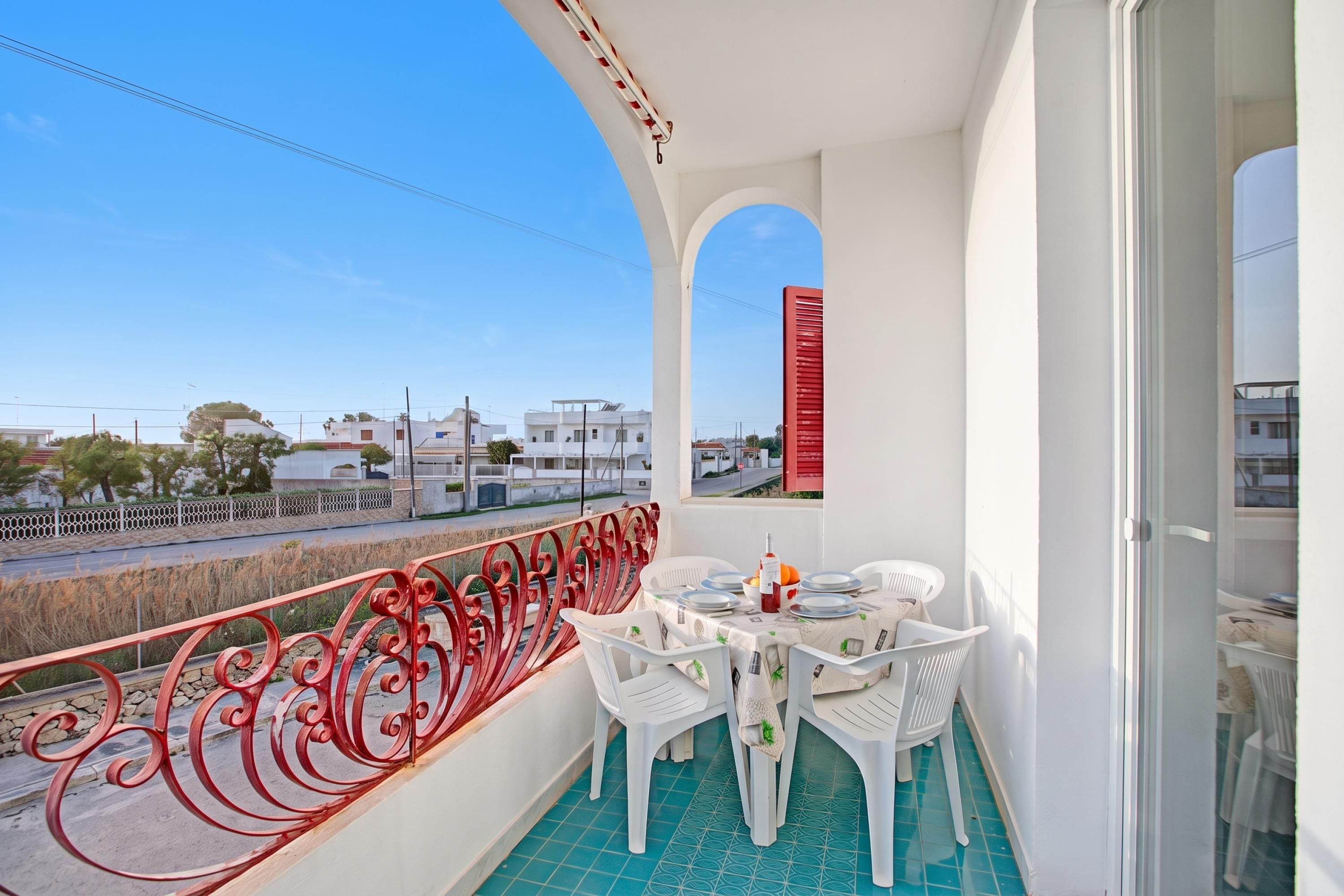 A balcony with a table set for dining, a red railing, and a view of the street and buildings.