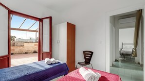 Une chambre avec deux lits, l’un recouvert d’une couverture bleue et l’autre d’une couverture rouge, une armoire en bois, une chaise et une vue sur l’extérieur.
