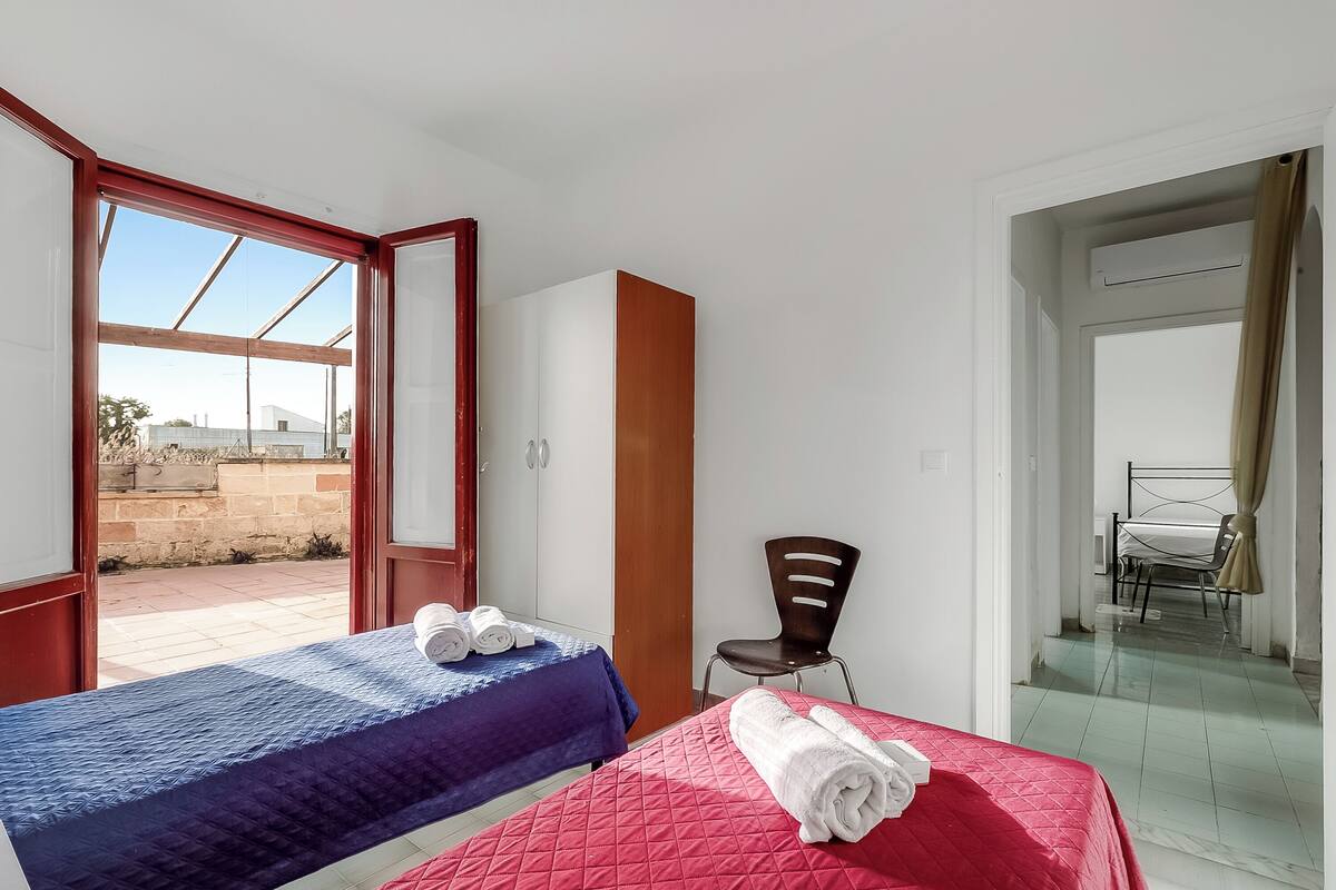 Une chambre avec deux lits, l’un recouvert d’une couverture bleue et l’autre d’une couverture rouge, une armoire en bois, une chaise et une vue sur l’extérieur.