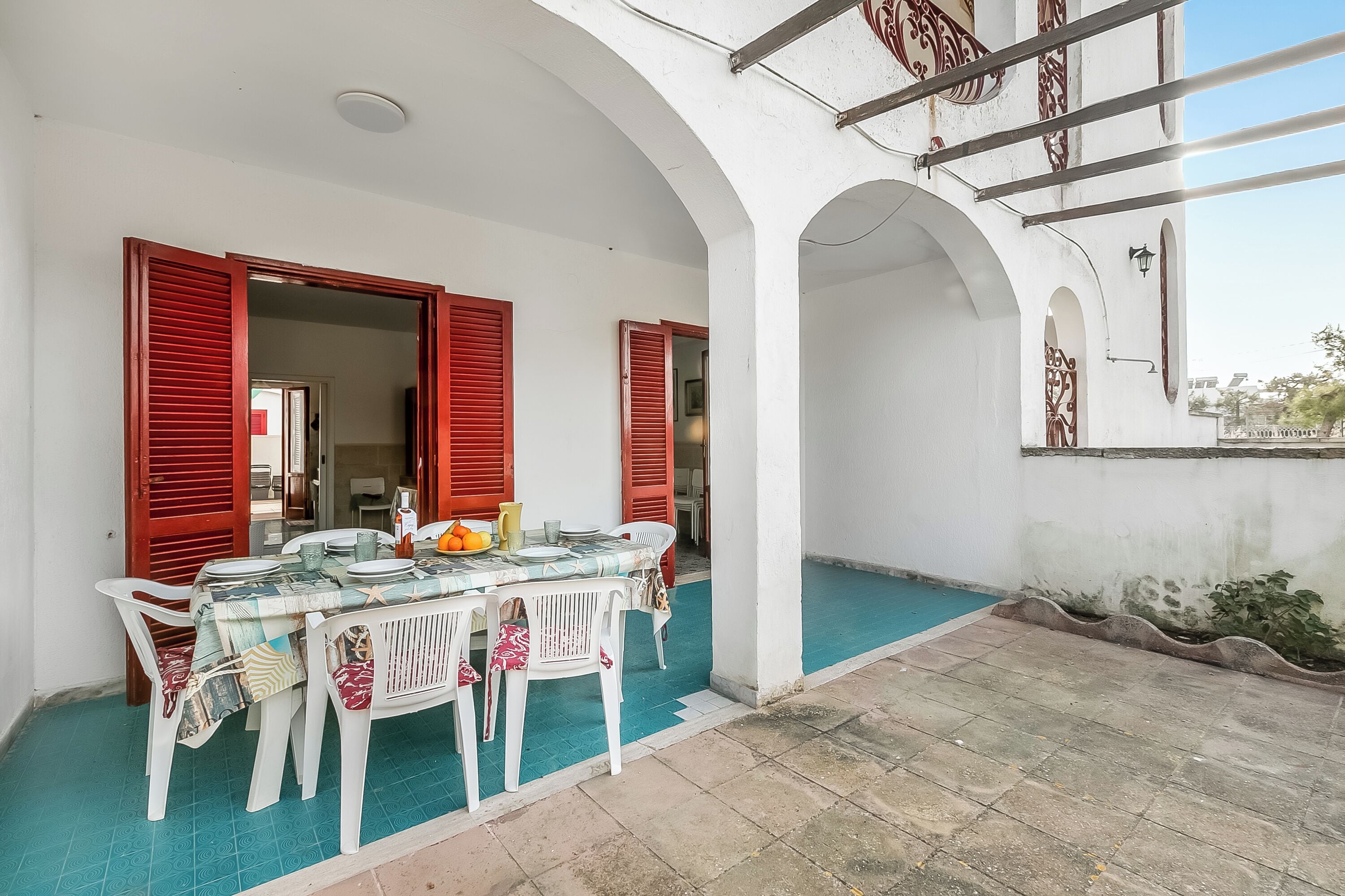 An outdoor dining area with a table set for four, surrounded by white plastic chairs, under a white building with red shutters.