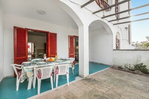 An outdoor dining area with a table set for four, surrounded by white plastic chairs, under a white building with red shutters. - Residence Vista Mare (Porto Cesareo)