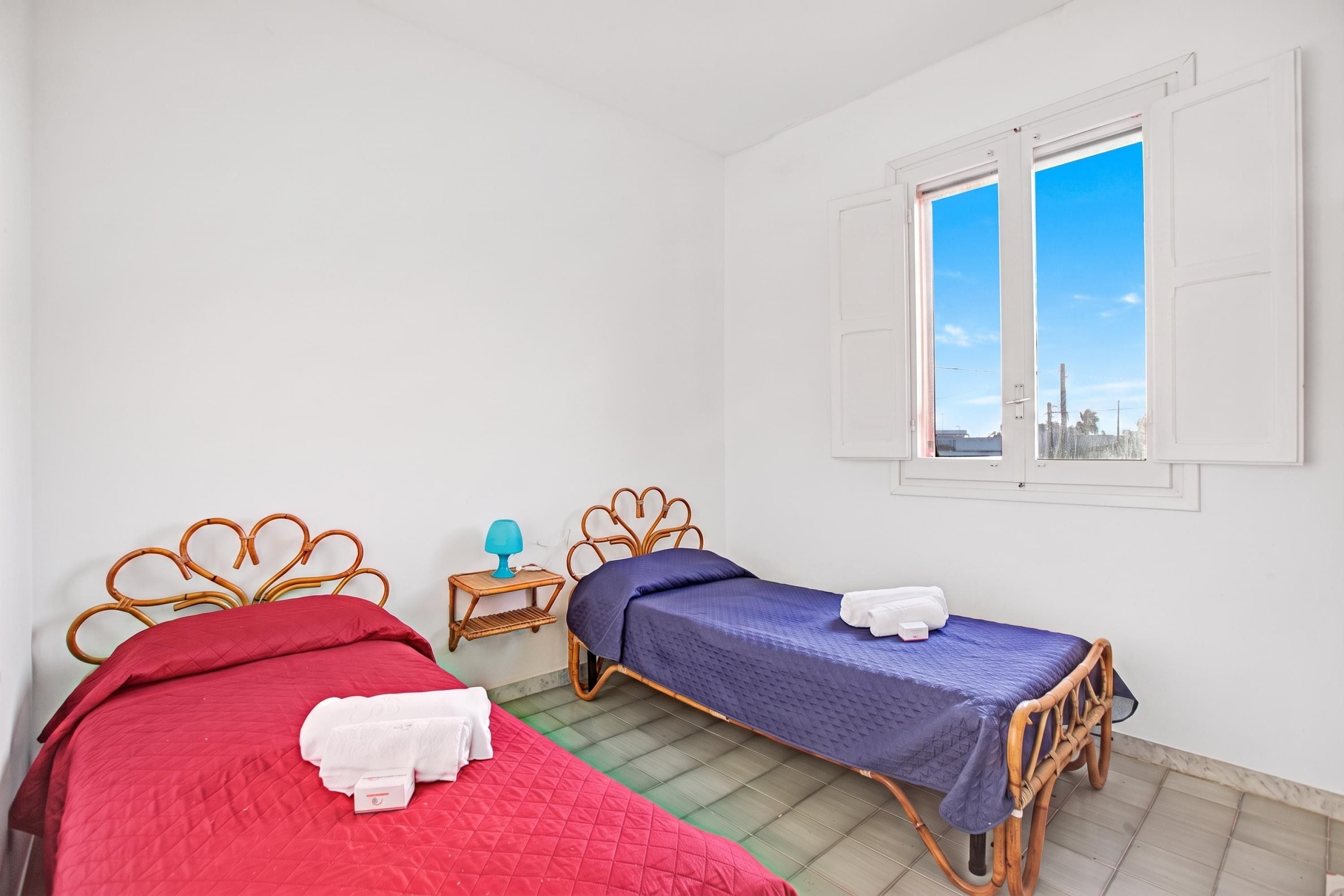 A room with two beds, a window with shutters, a small table with a lamp, and a view of the city.