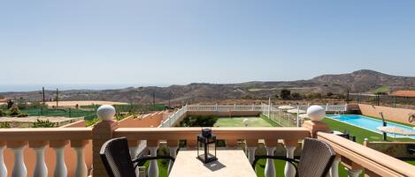 A balcony with two chairs and a table, overlooking a pool and a hilly landscape.
