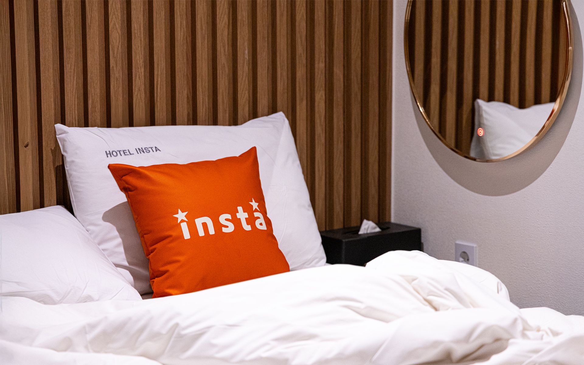 A hotel room with a bed, a pillow with the logo 'insta', a bedside table, and a round mirror on the wall.