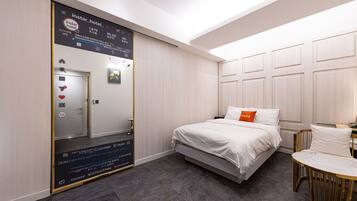 Habitación de hotel con cama, mesita de noche, silla, espejo y pantalla digital en la pared.