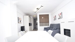 Een moderne, goed verlichte woonkamer met een witte eettafel, een grijze bank en een televisie op een dressoir.