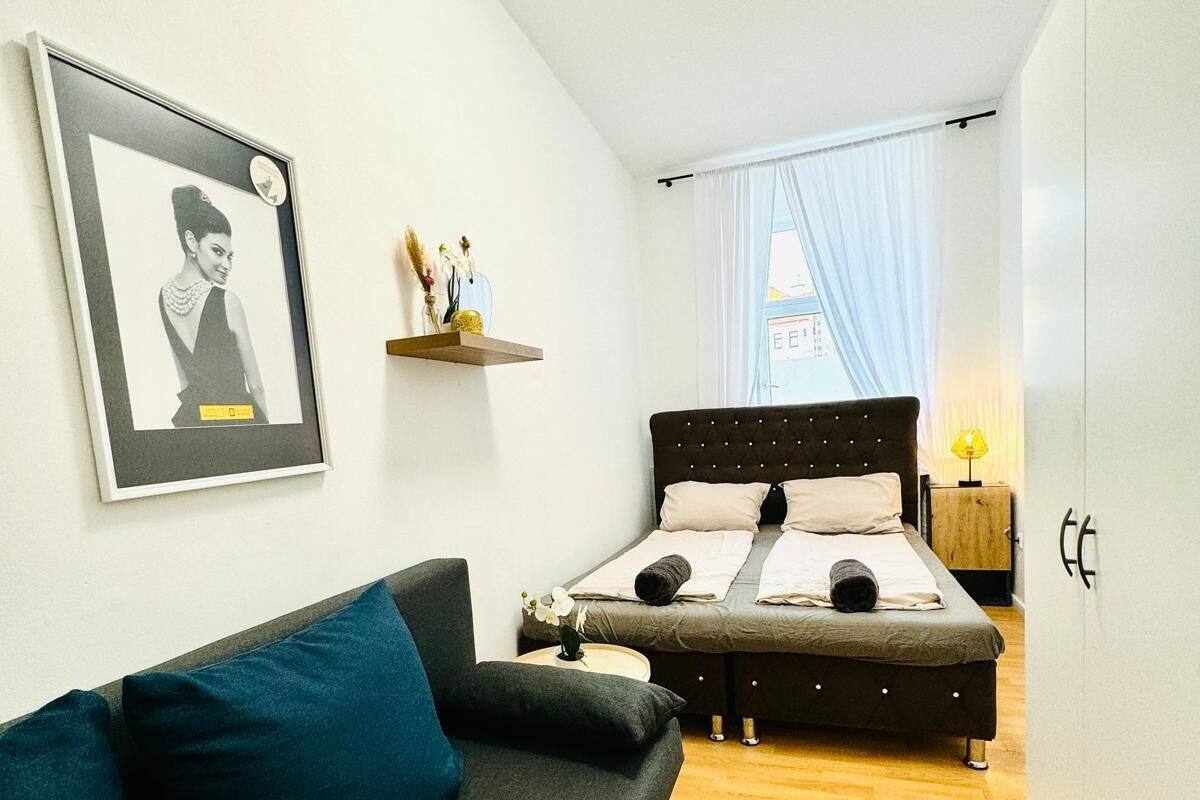 A compact bedroom with a bed, a sofa, a small bedside table, a nightstand, and a window with curtains.