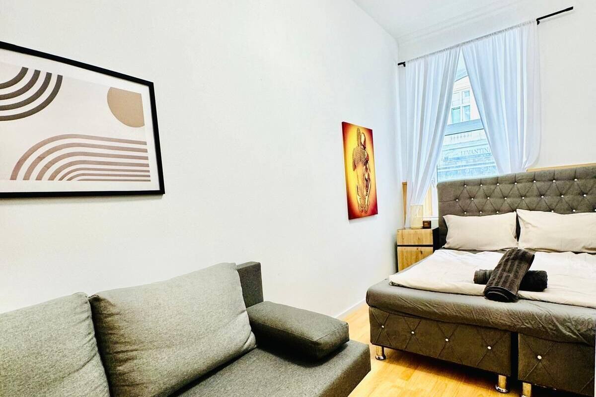 A modern bedroom with a grey sofa, a bed with a tufted headboard, and a framed abstract painting on the wall.
